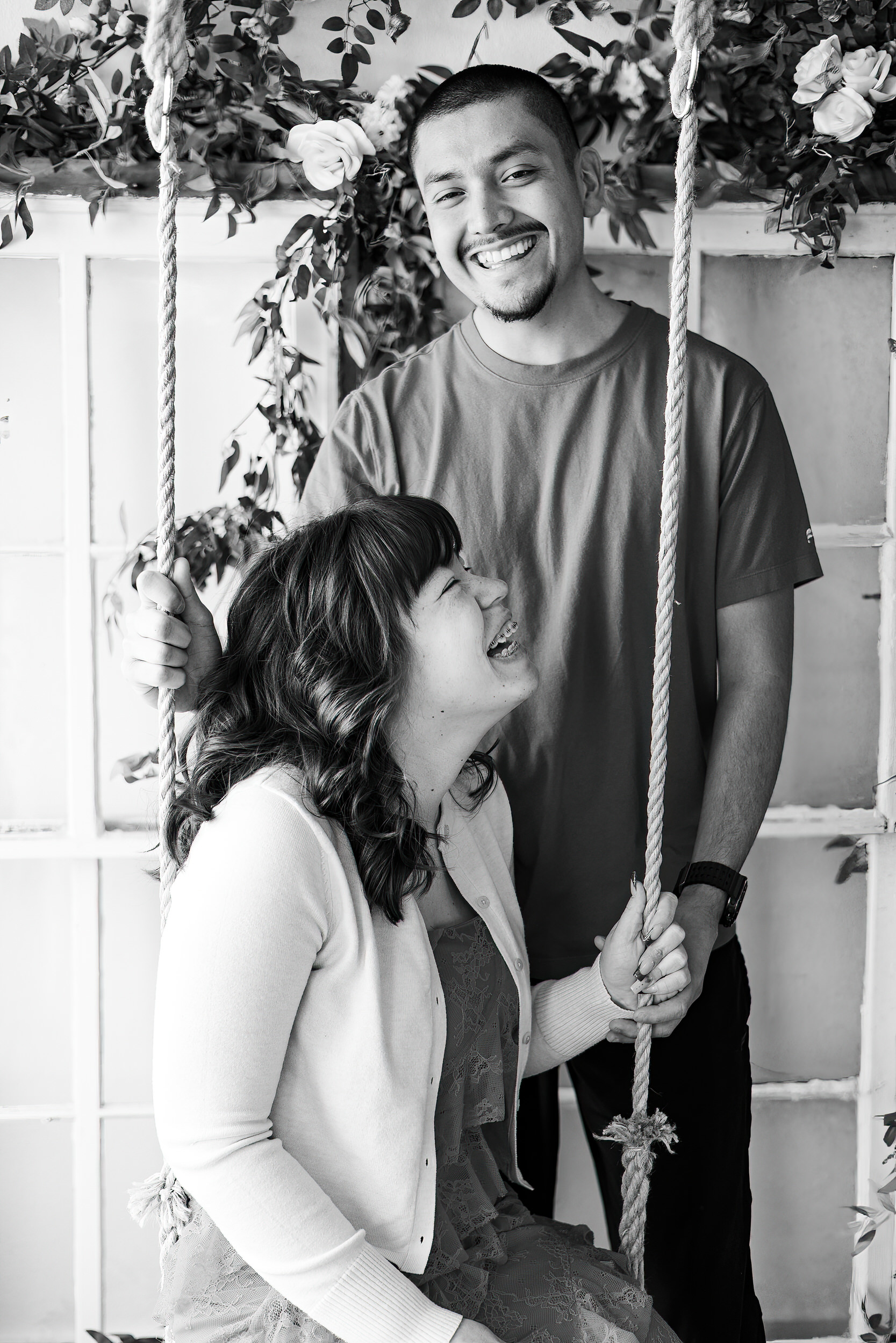 A woman sits on a wooden swing and her husband stands behind her as they laugh together.