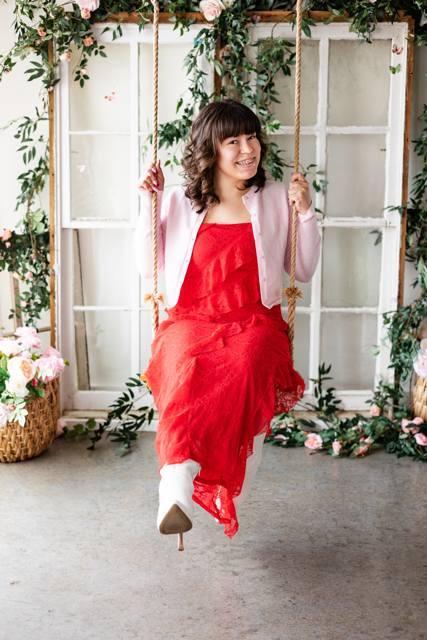 A woman in a red dress sits on wooden swing and smiles at the camera.