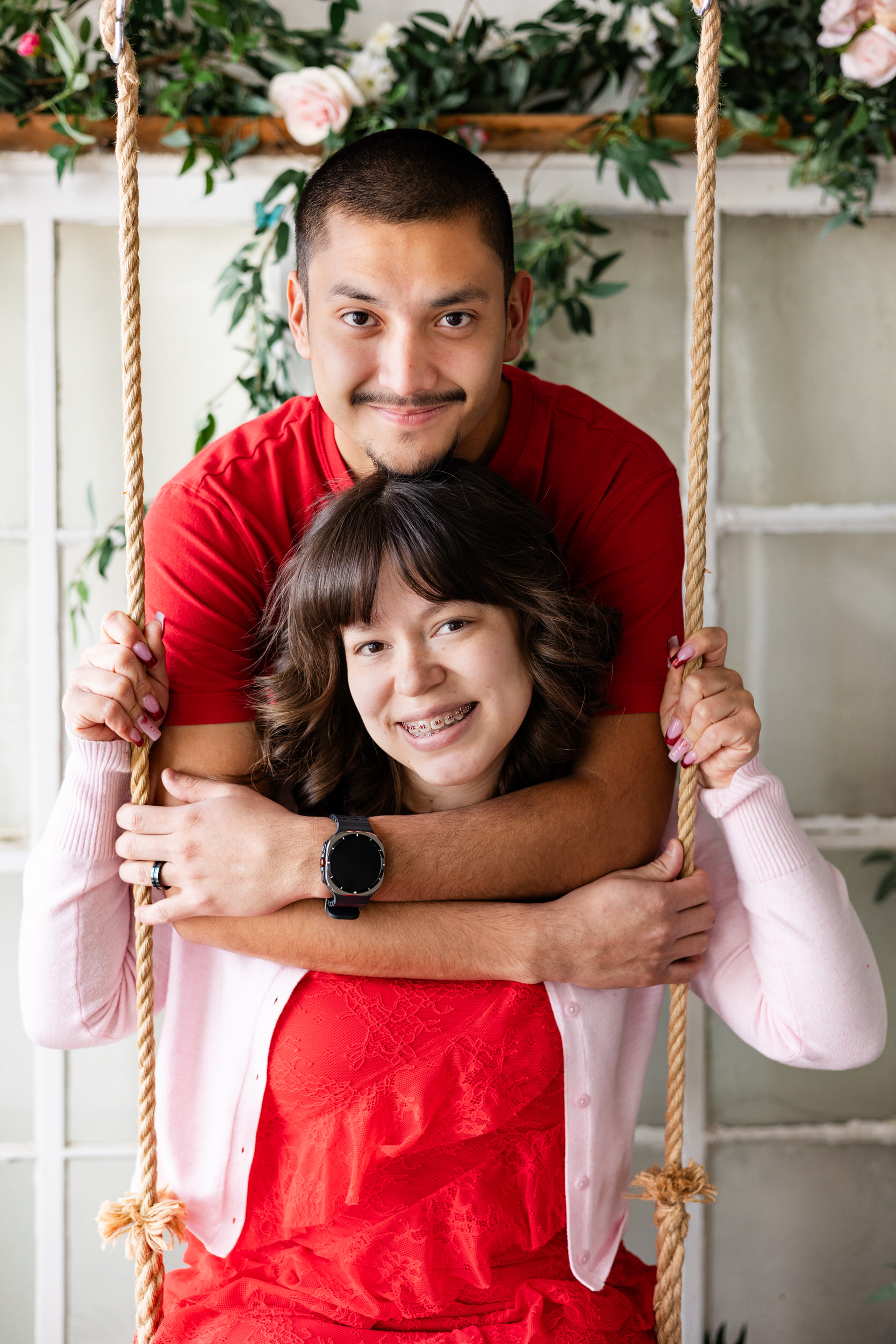 A woman sits on a wooden swing and her husband hugs her around the shoulders from behind and they both smile at the camera.