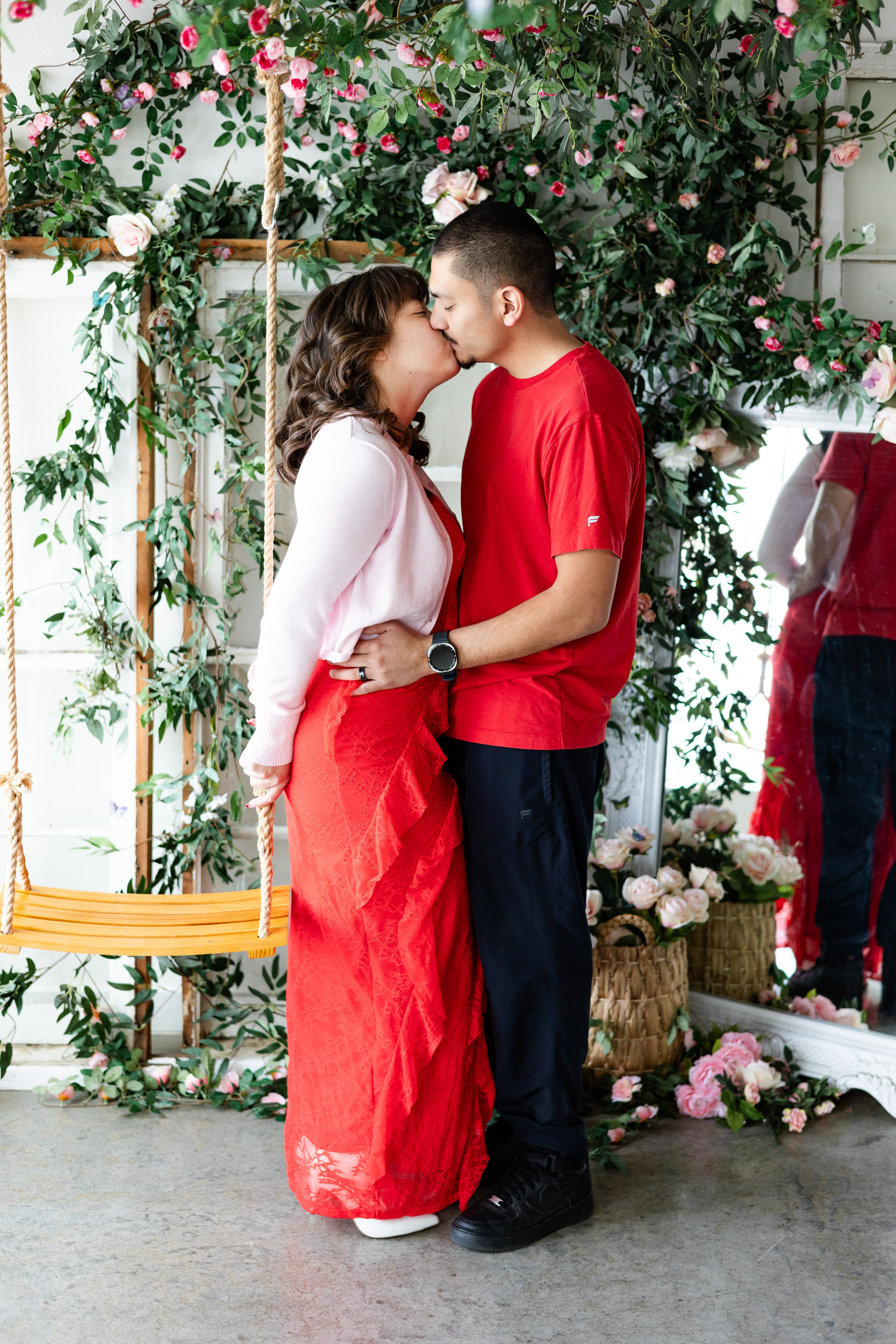 A woman stands with her back to the rope of a wooden swing and shares a kiss with her husband.