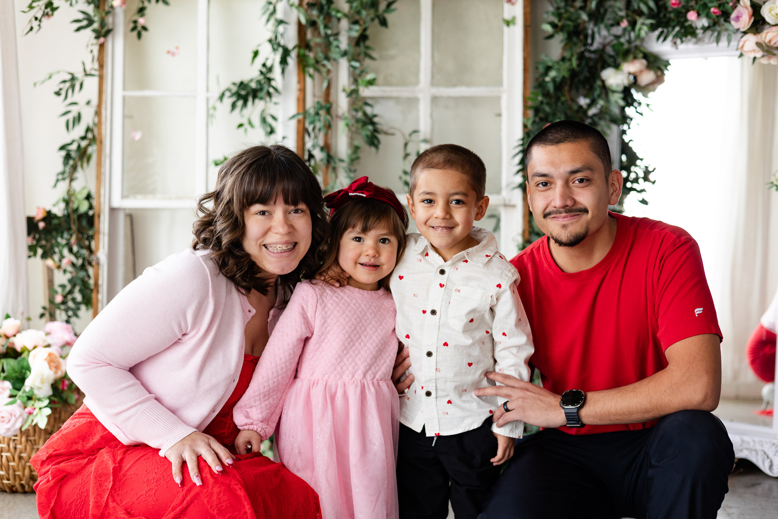 A mom and dad squat down next to their young son and daughter and they all smile at the camera.