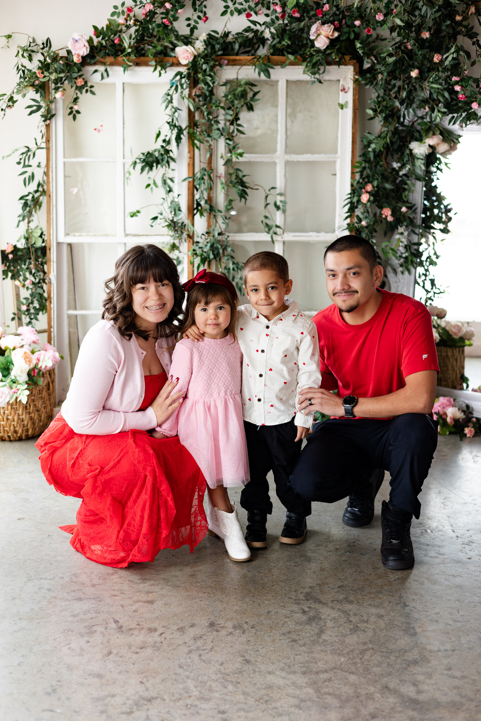 A mom and dad squat down next to their young son and daughter and they all smile at the camera.