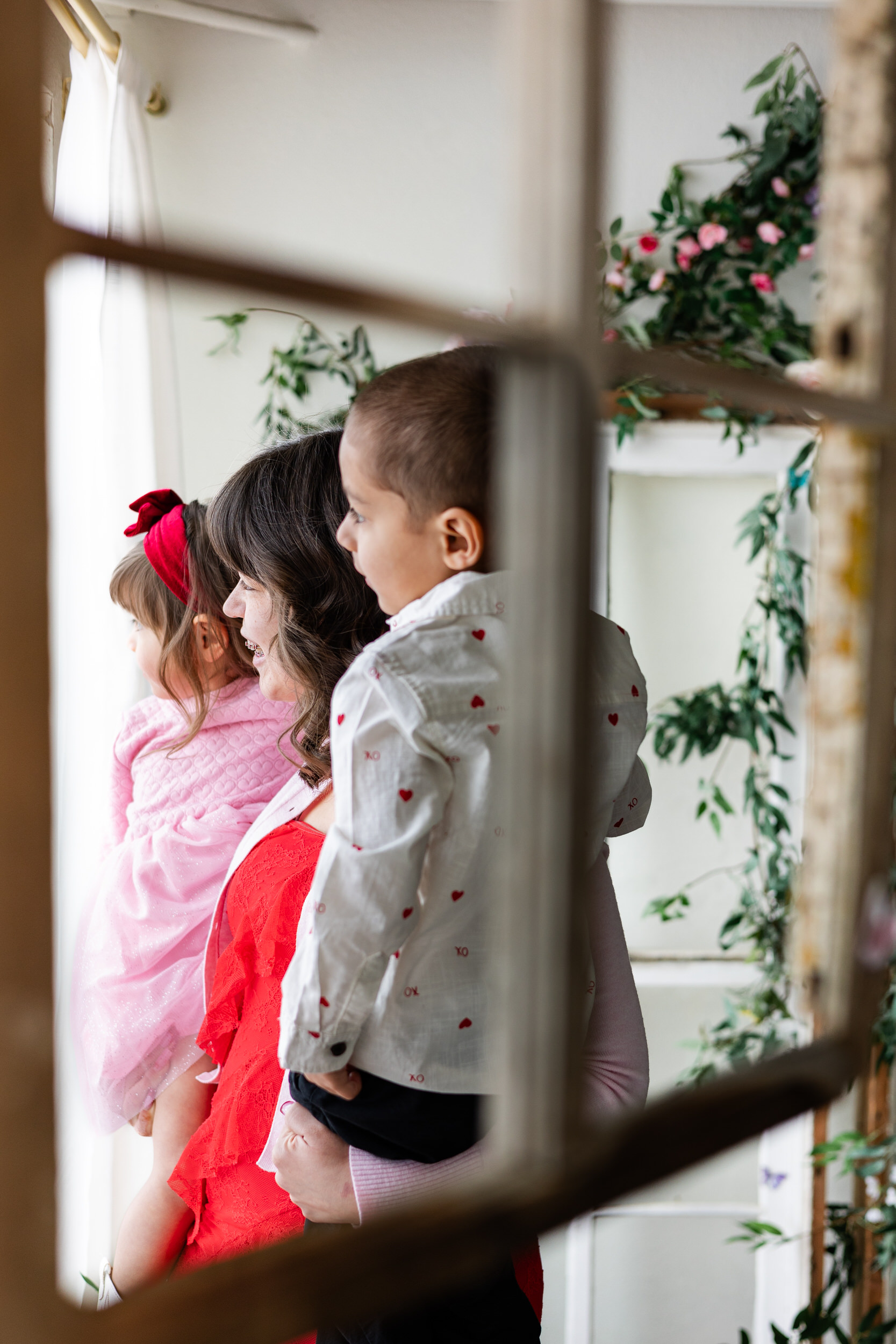 A mom holds her young son and daughter on her hips and they look out a window.