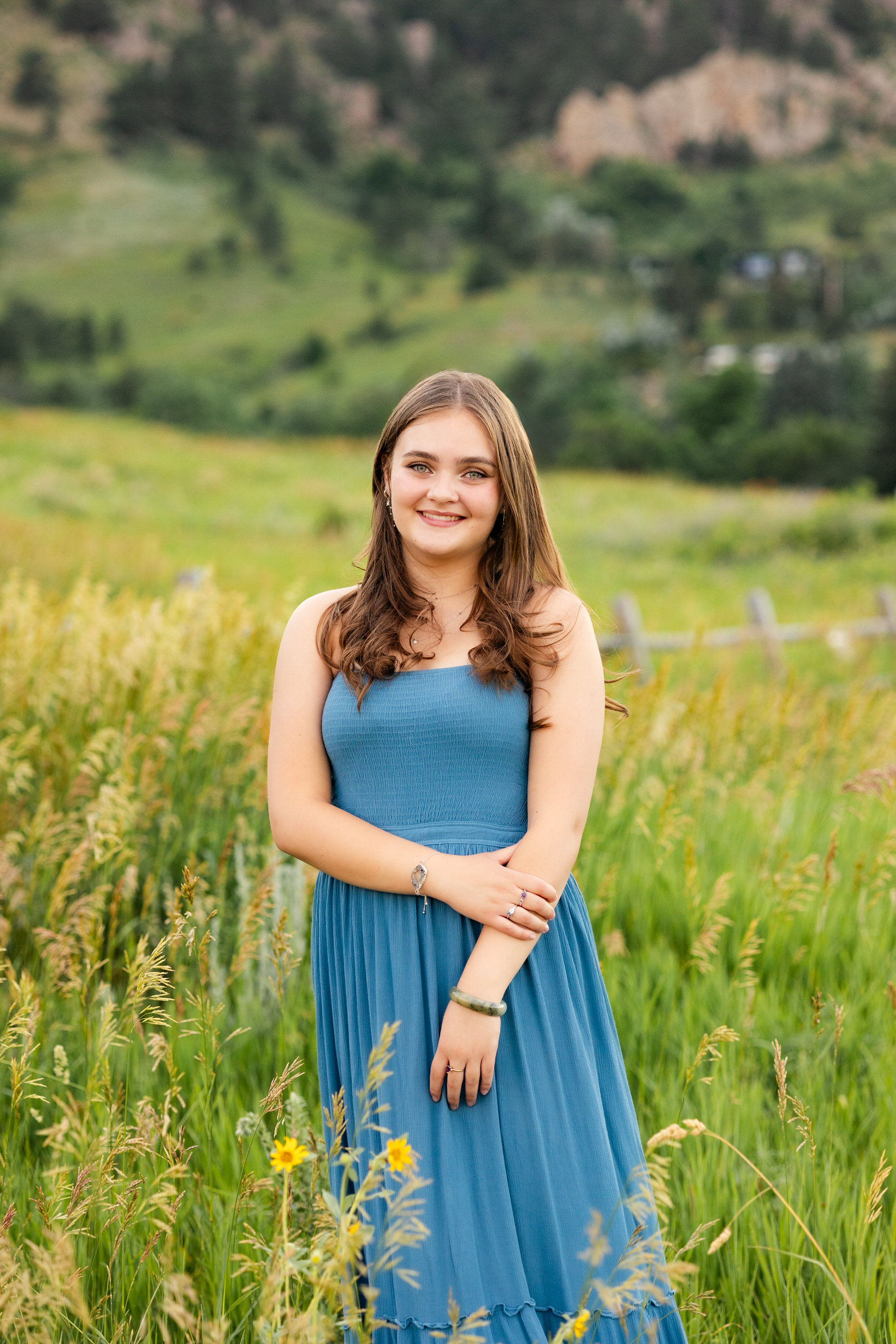 A woman in a teal dress stands in a field and smiles a the camera.