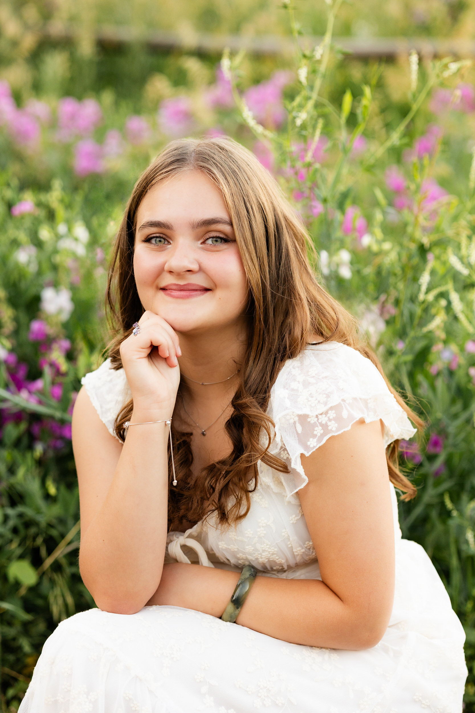 A woman in a white dress squats in a field of wildflowers and smiles at the camera.