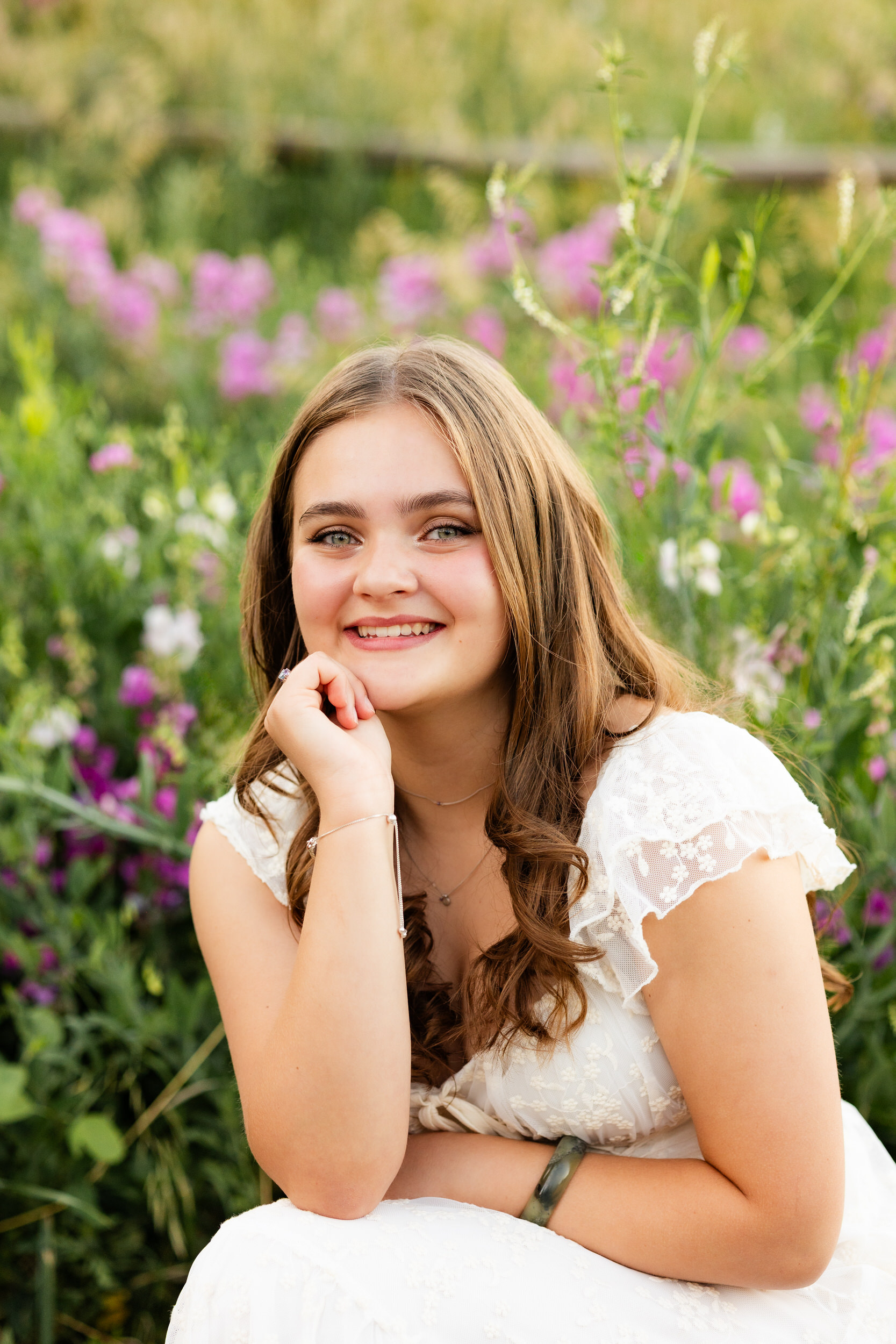 A woman in a white dress squats in a field of wildflowers and smiles at the camera.