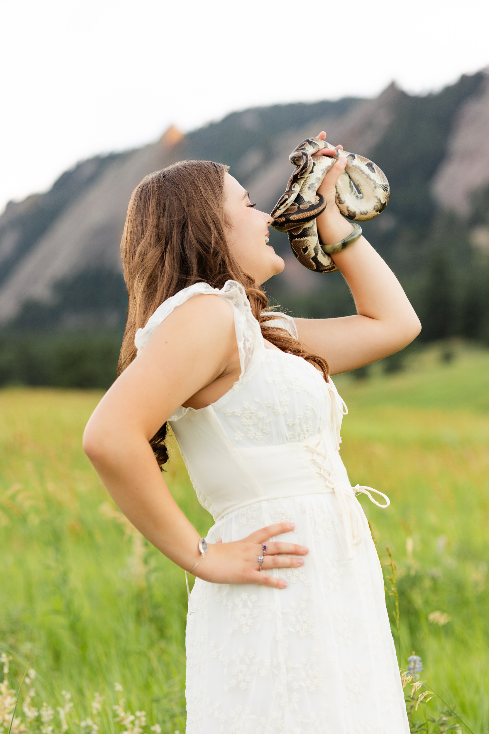 A woman in a white dress stands in a field holding her snake and the snake "kisses" her nose.