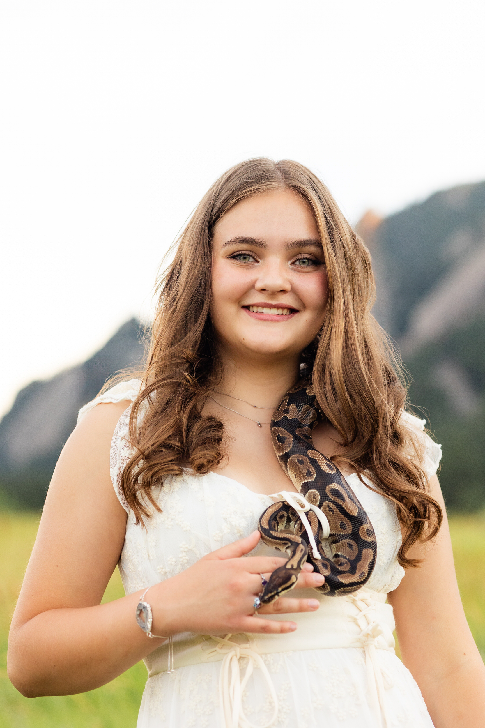 A woman in a white dress with a snake on her shoulder smiles at the camera.