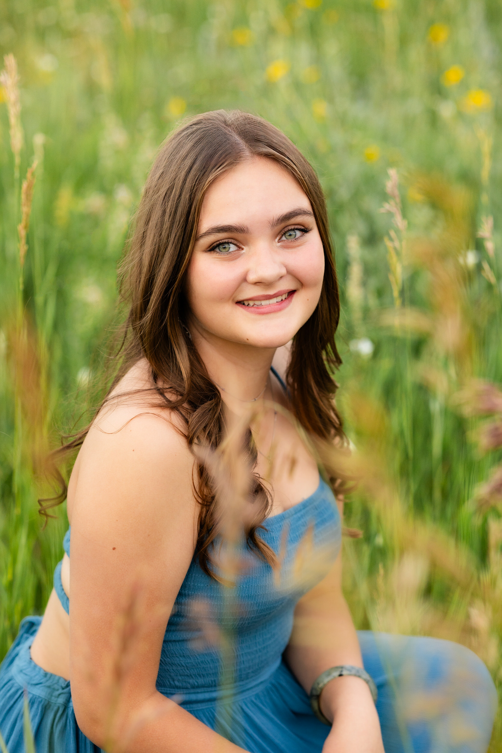 A woman in a teal dress squats in a field and smiles at the camera.