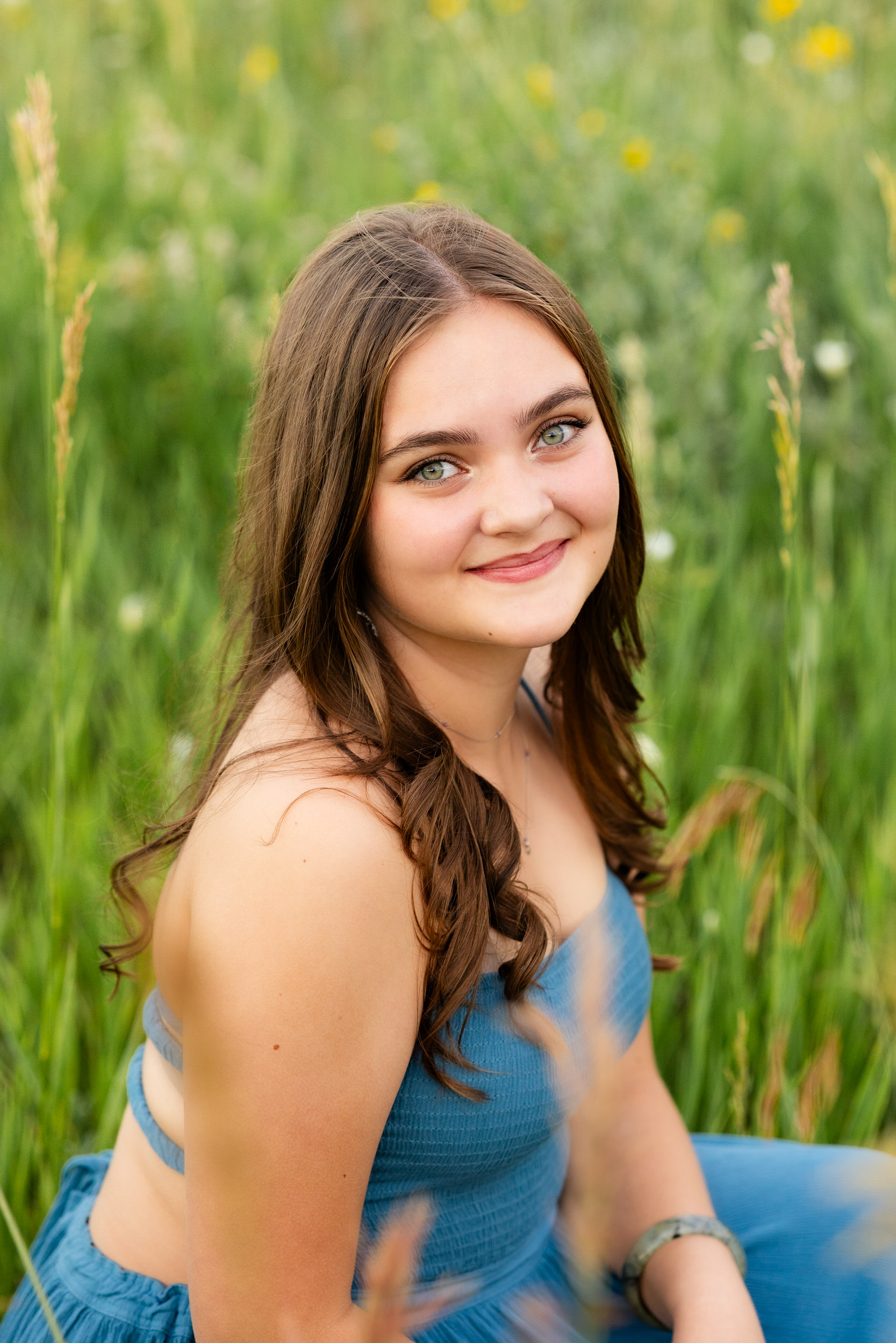 A woman in a teal dress squats in a field and smiles at the camera.