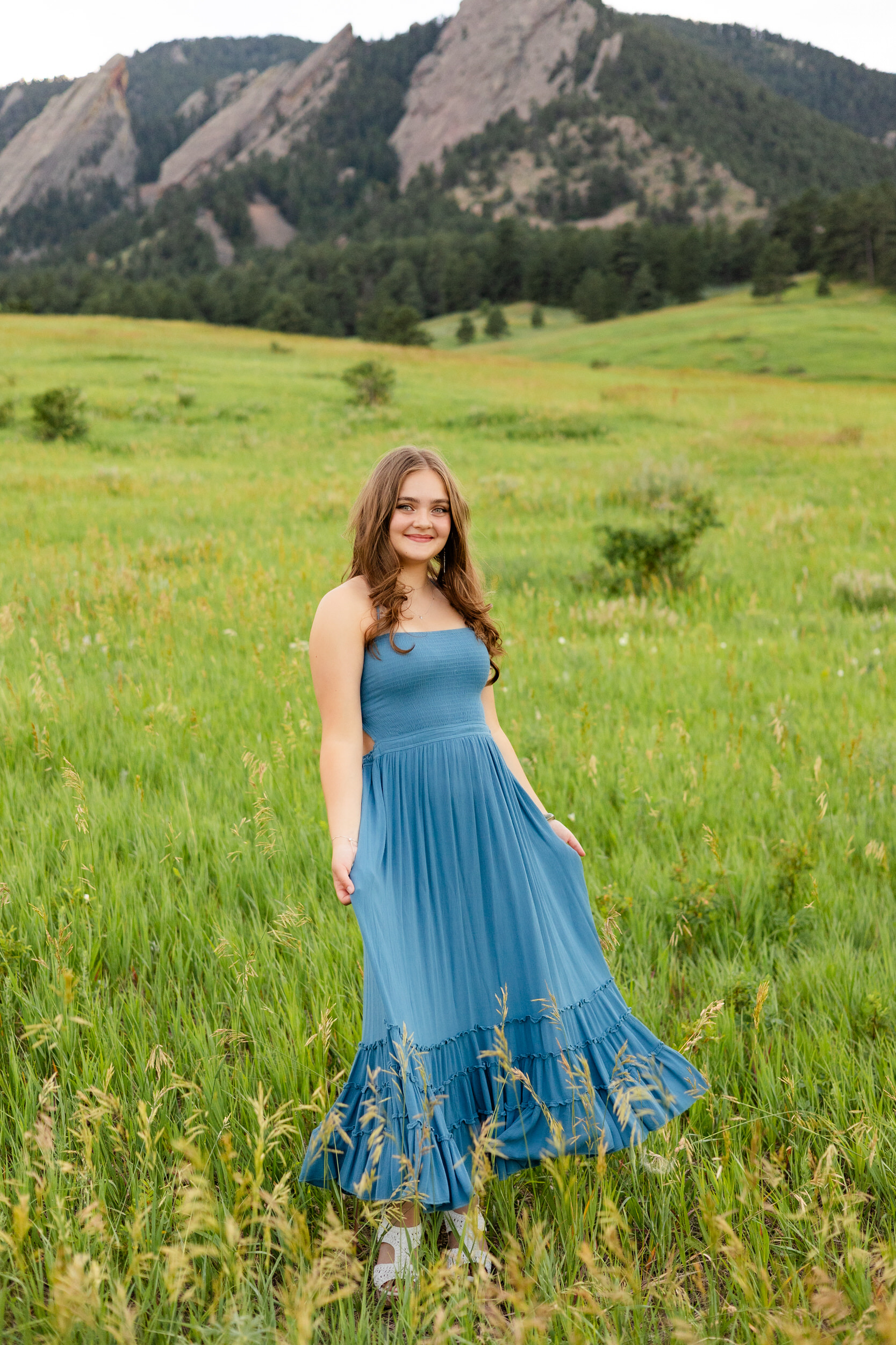 A woman in a teal dress swishes her skirt in a field and smiles at the camera.