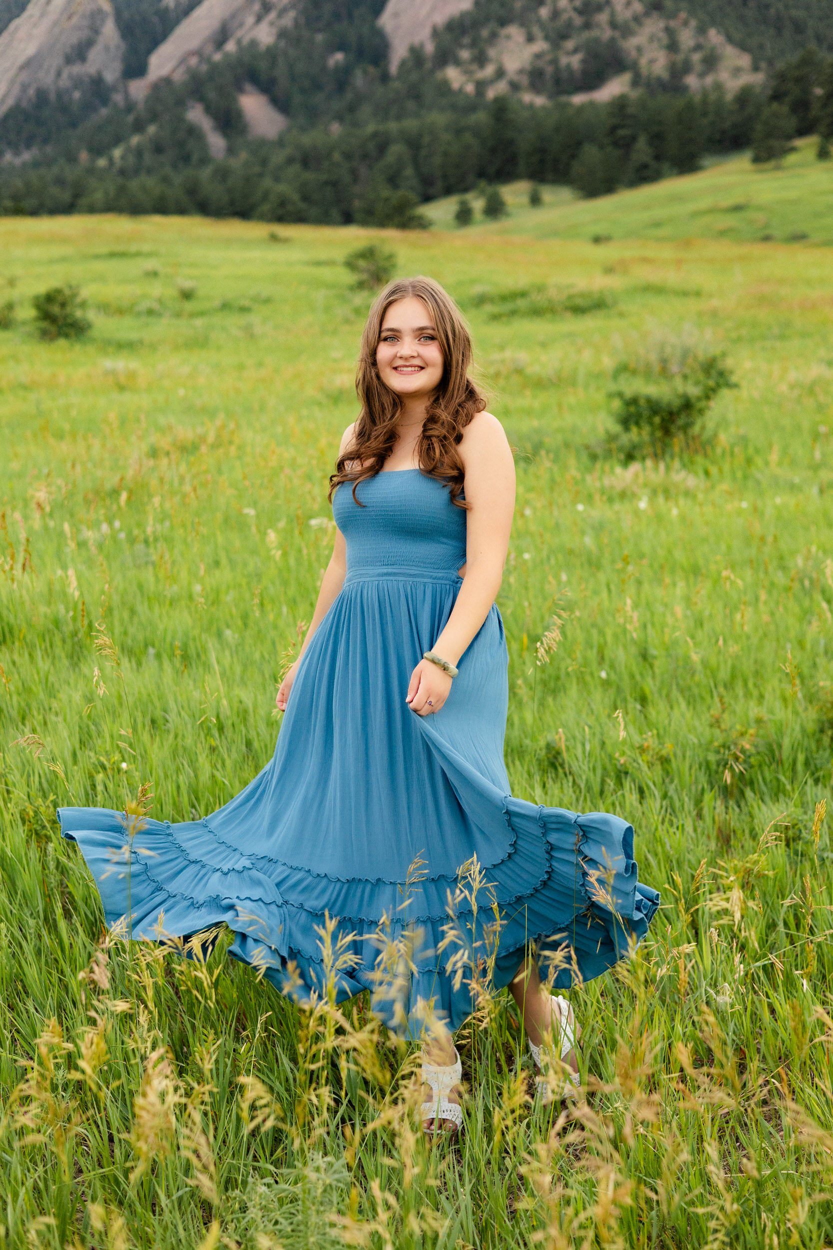 A woman in a teal dress swishes her skirt in a field and smiles at the camera.
