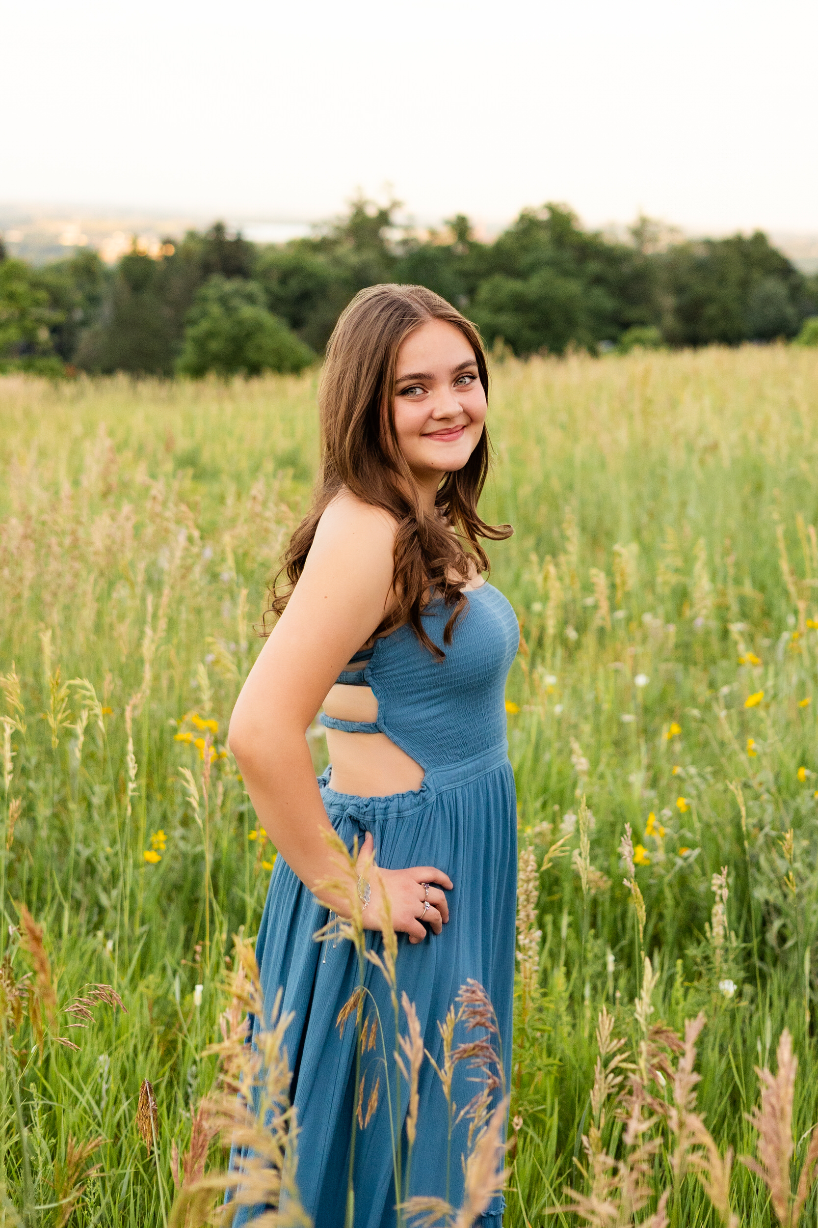 A woman in a teal dress stands in a field and smiles at the camera over her shoulder.