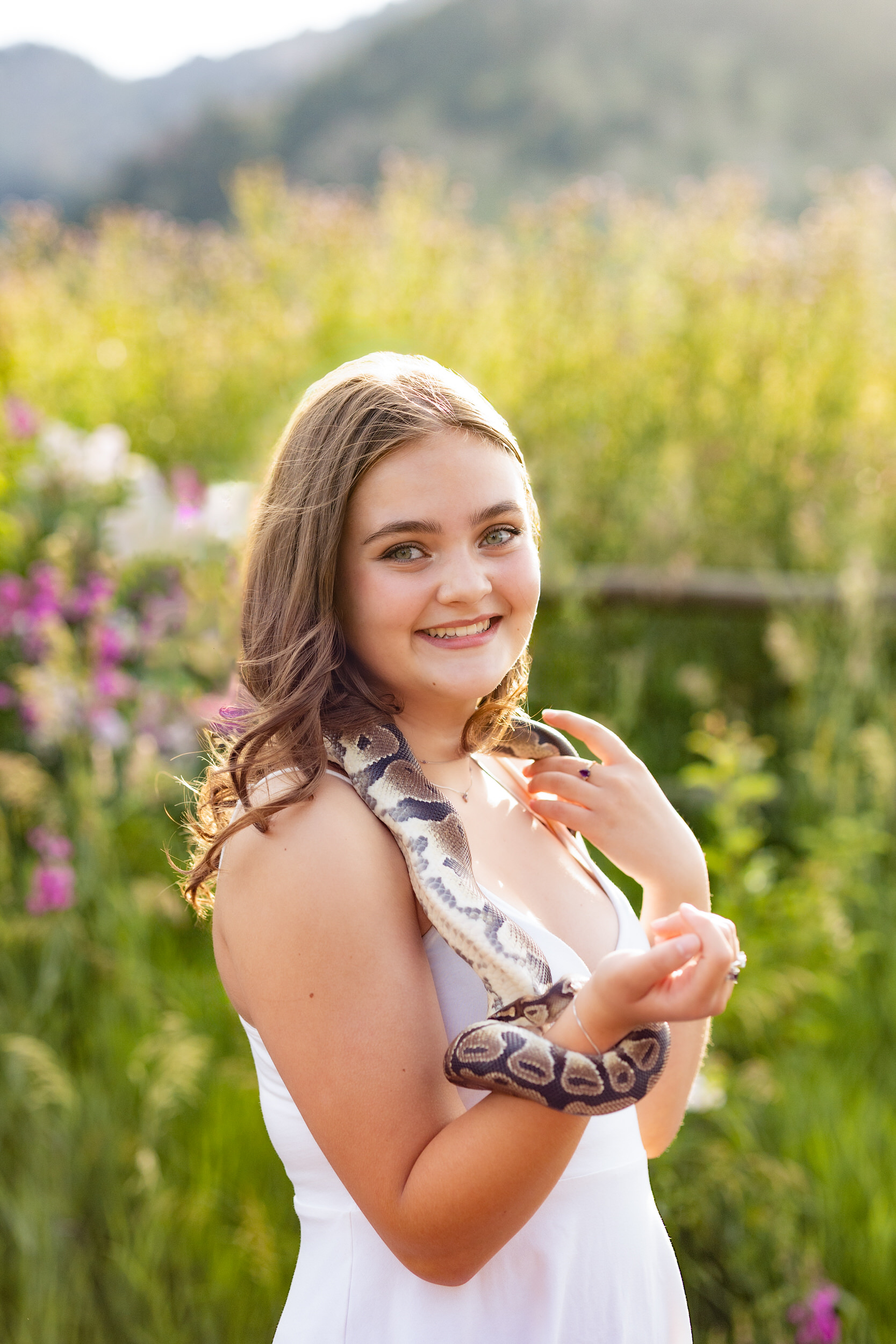 A woman in a white dress surrounded by wildflowers smiles at the camera with a snake wrapped around her arm.