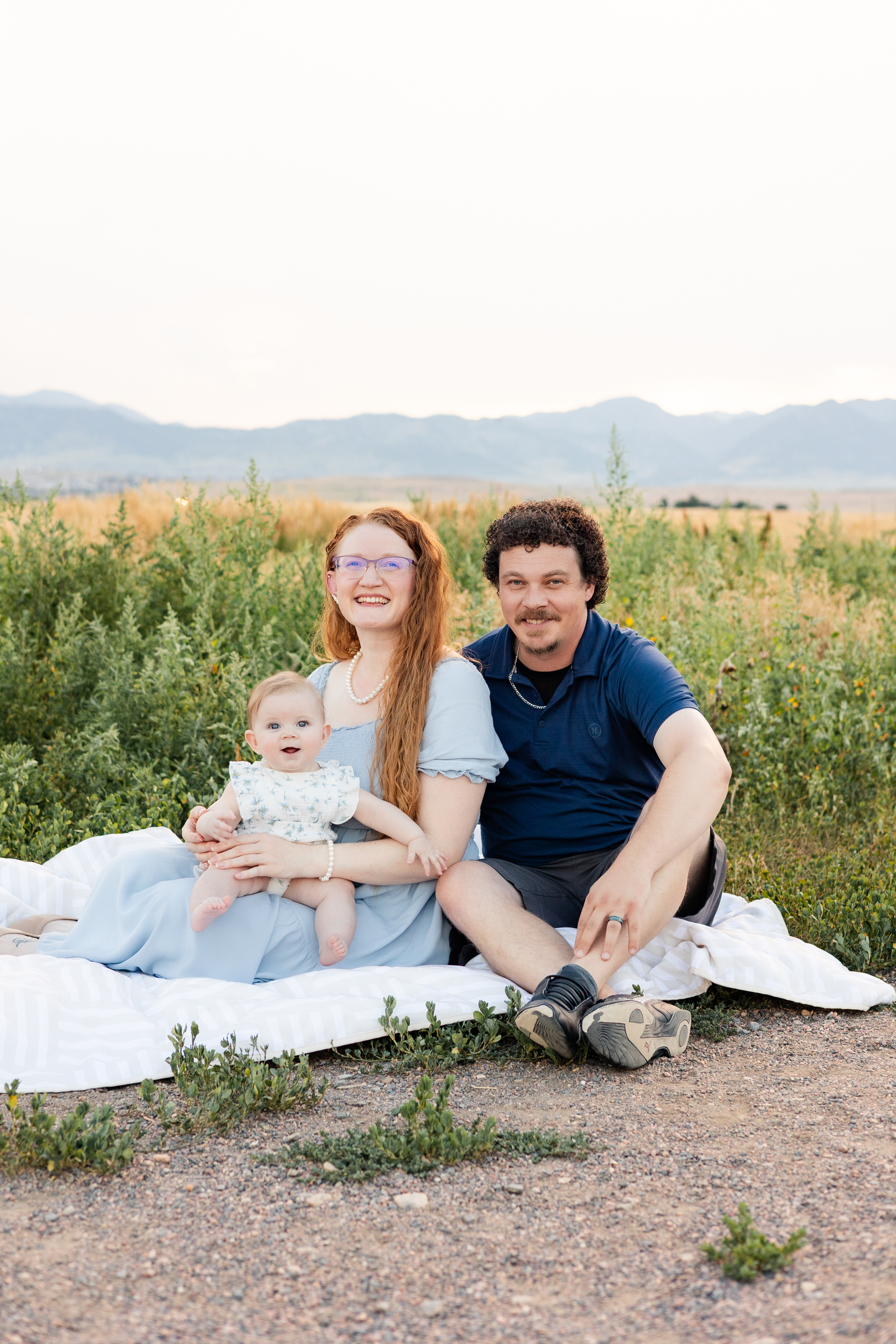 Mom and dad sit on blanket while baby girl sits in mom's lap.