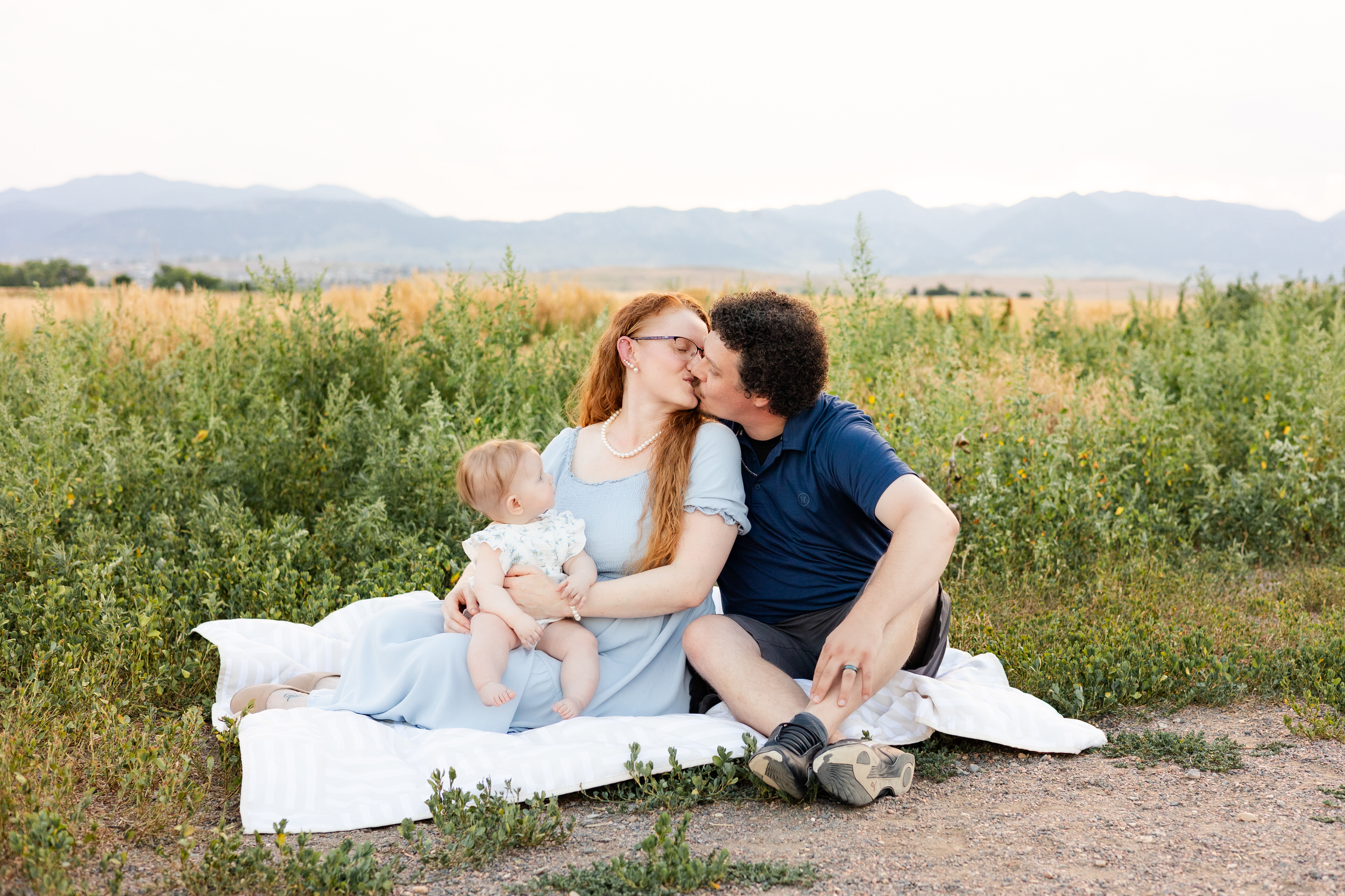 Mom and dad sit on a blanket and share a kiss while baby girl sits in mom's lap.