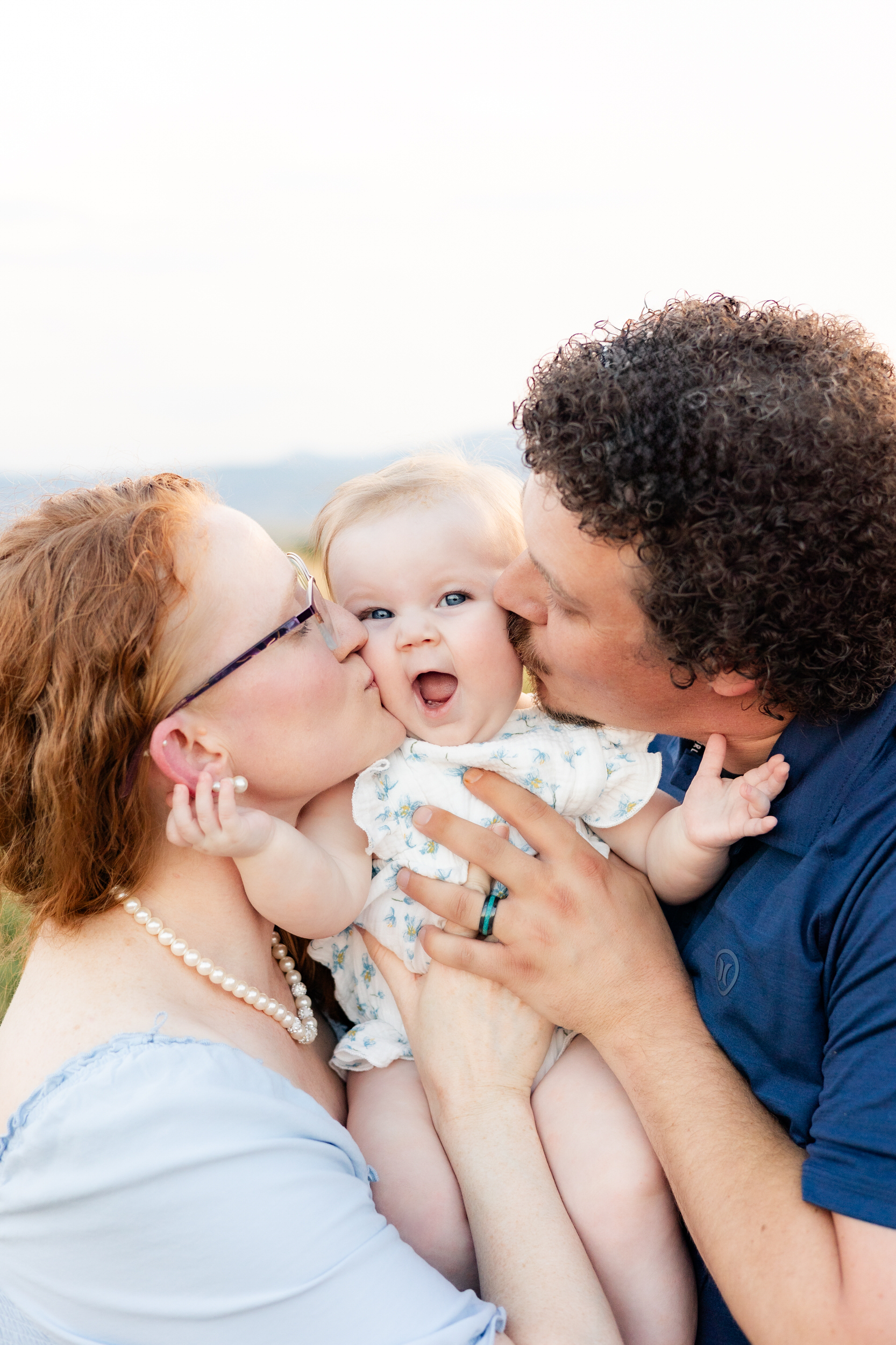 Mom and dad kiss their baby girl on her cheeks as she looks surprised.