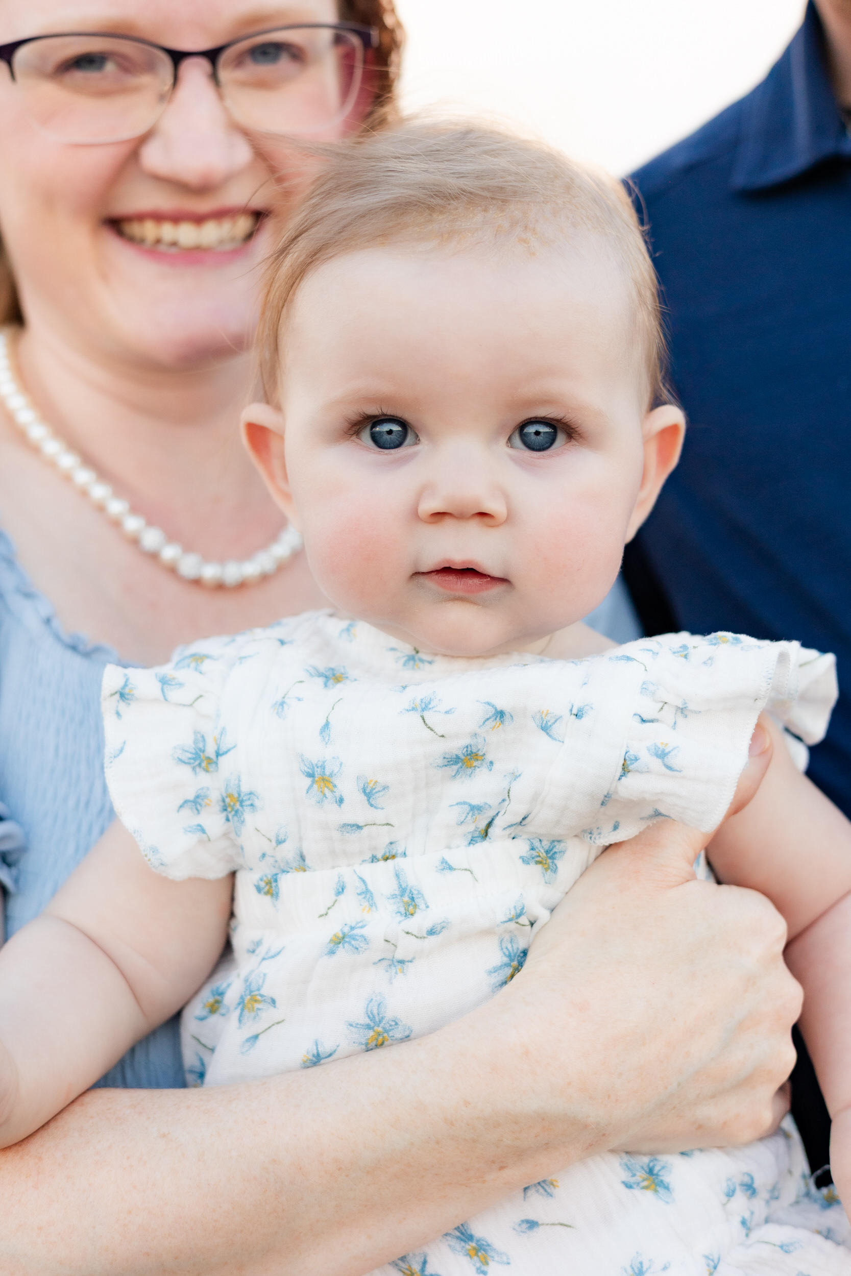 Baby girl held by her mom looks directly at the camera while mom smiles in the background.