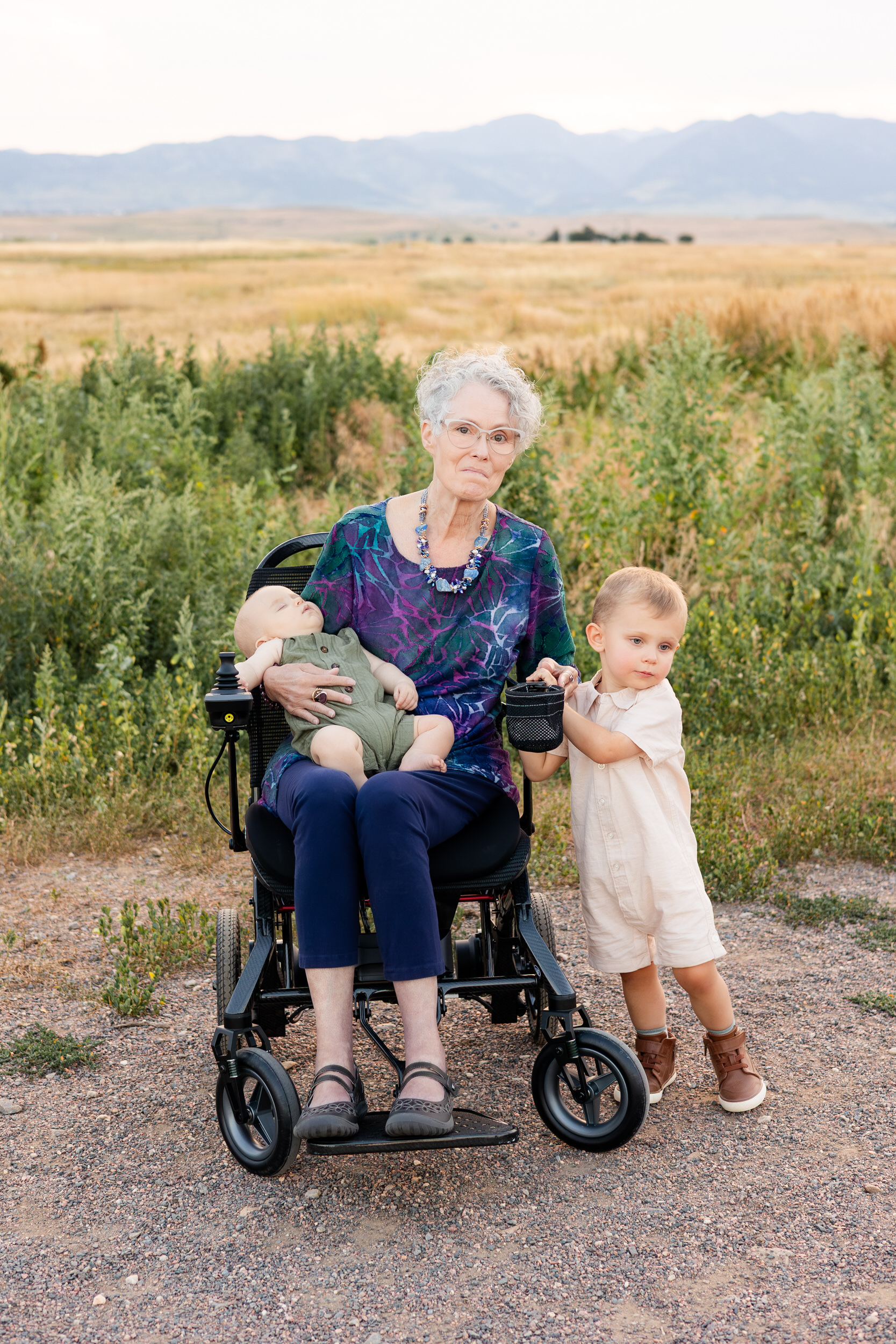 Grandma sits in wheelchair with grandbaby in her lap and grandson at her side.