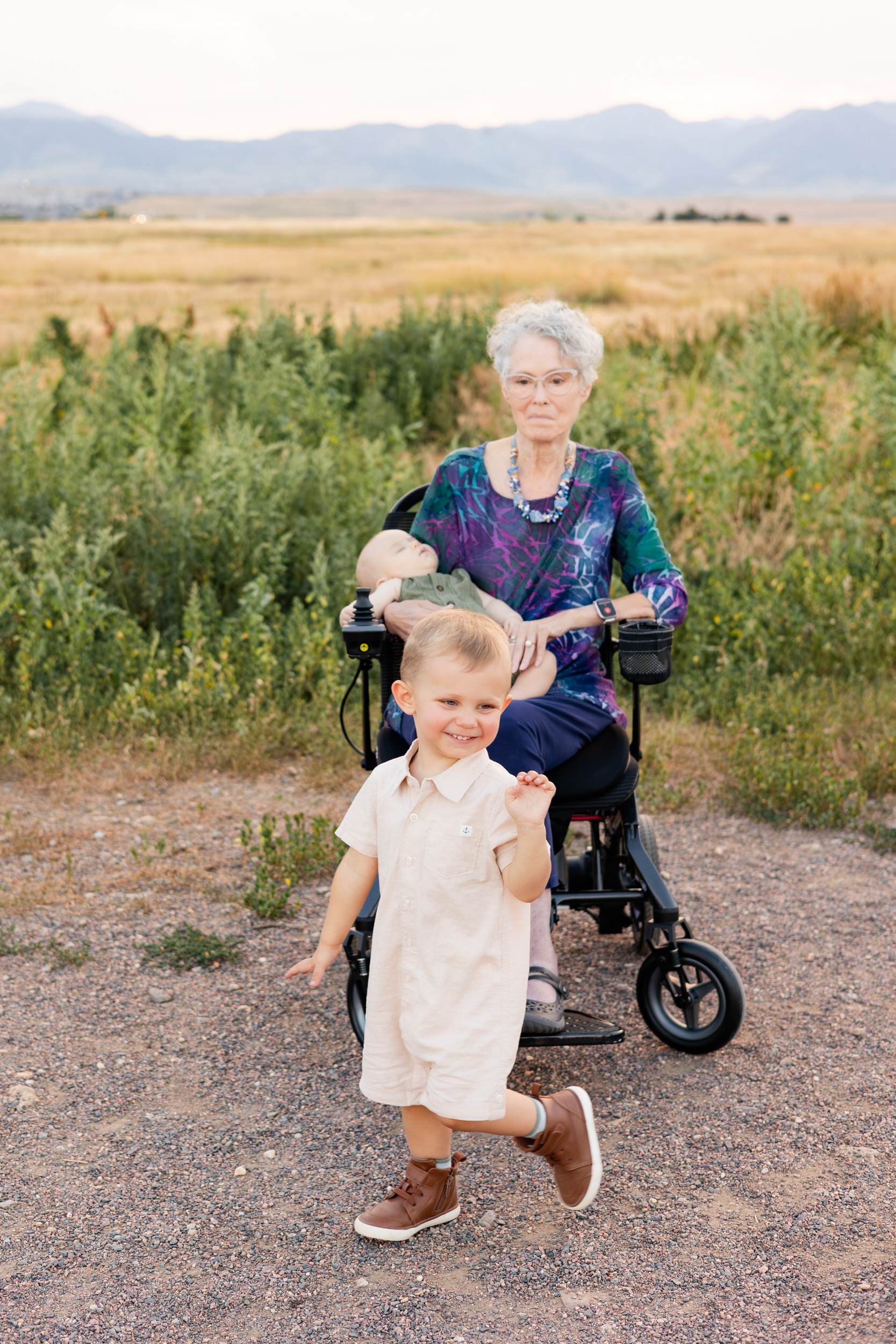 Grandma sits in wheelchair with grandbaby in her lap and grandson waving and smiling as he walks out of the picture.