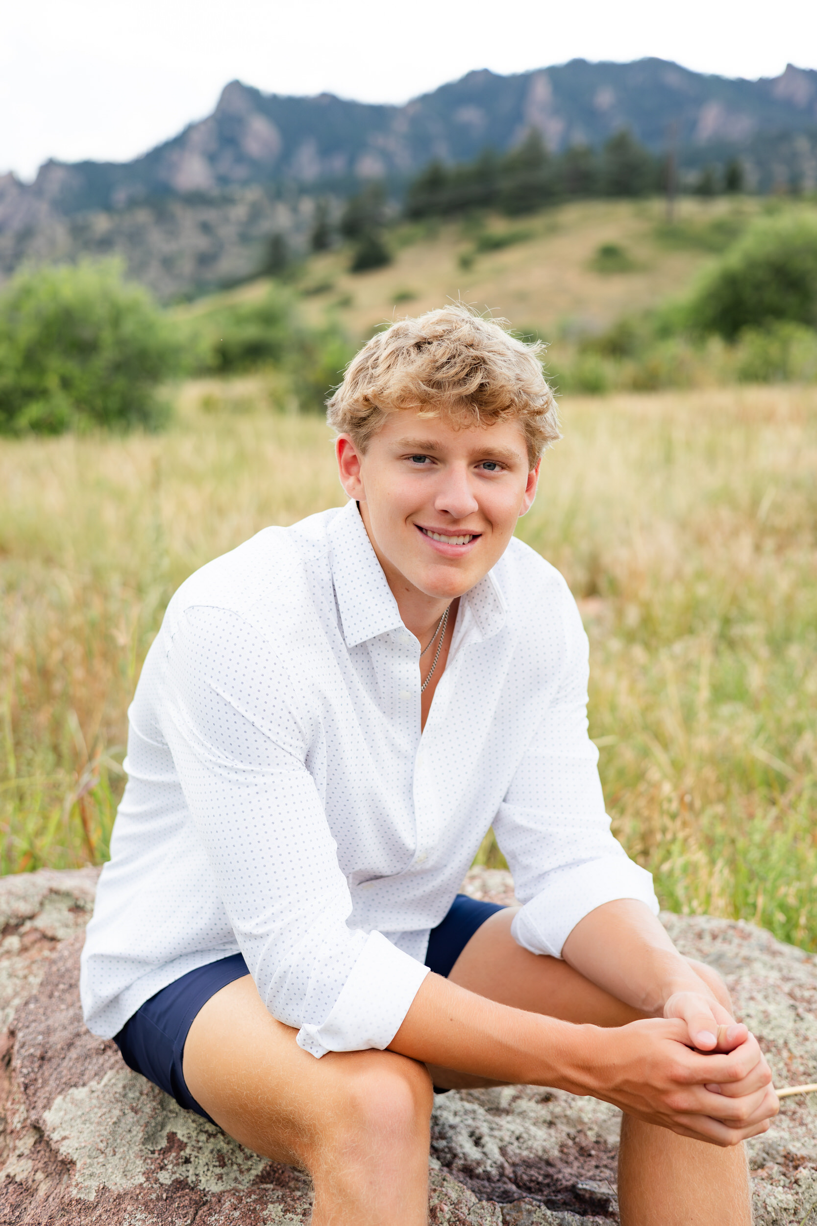 A young man sits on a boulder in a field and smiles at the camera.