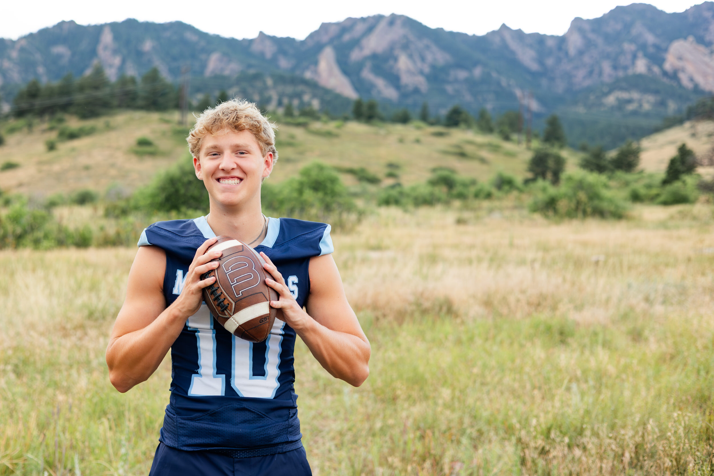 Young man in a school football jersey holds a football and stands in front of the mountains smiling.