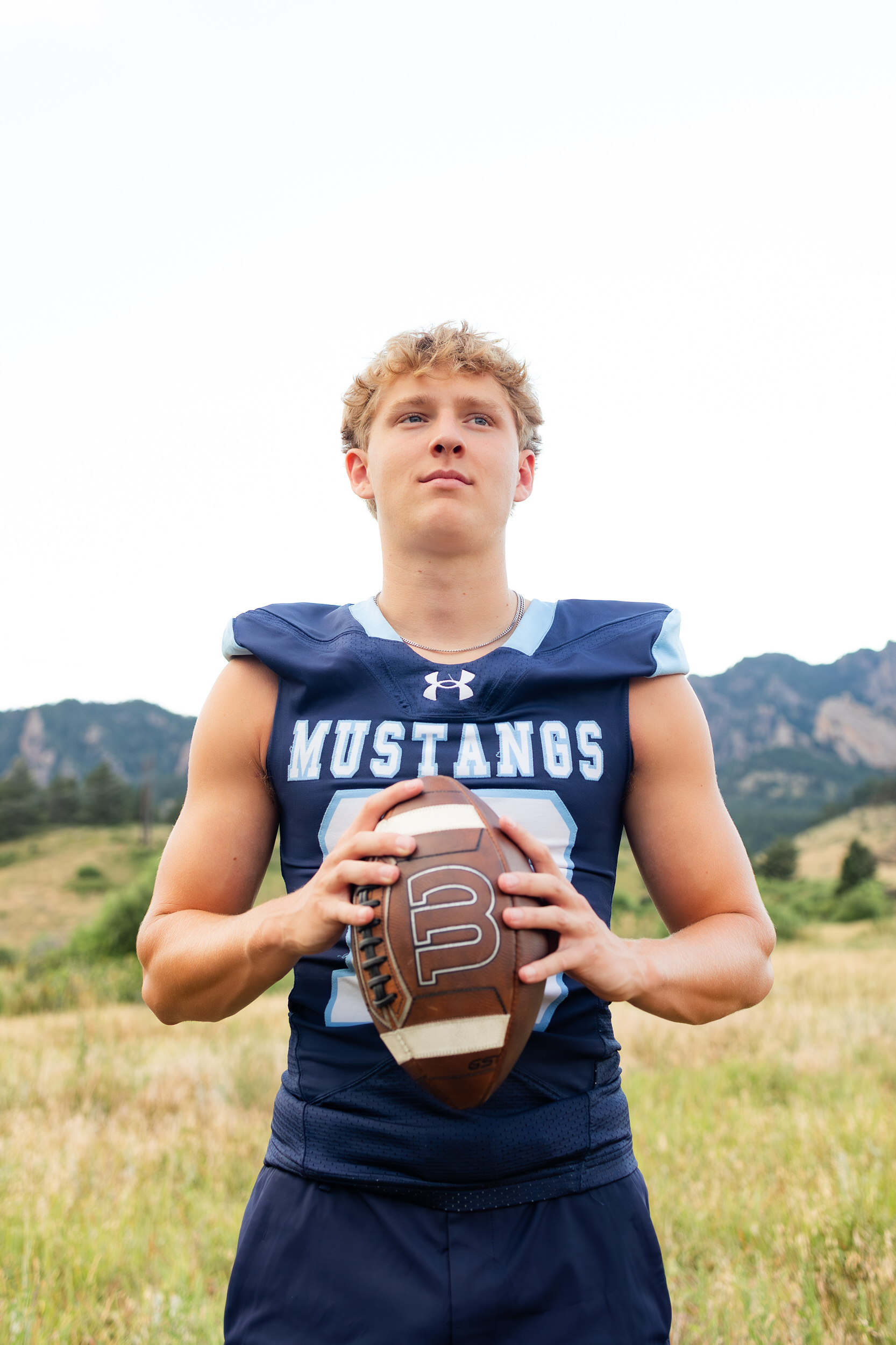 A young man stands in front of the mountains in a school football jersey and looks off camera.