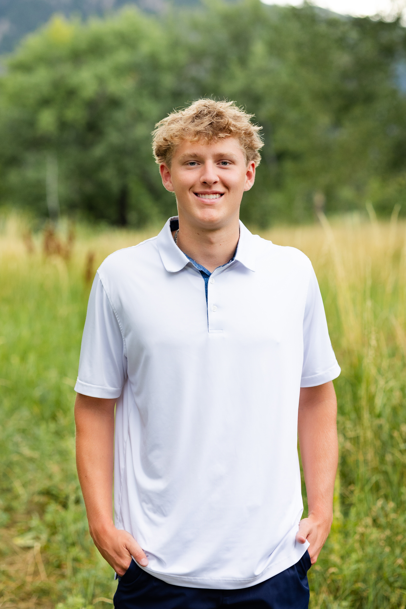 A young man stands in a field with his hands in his pockets and smiles at the camera.
