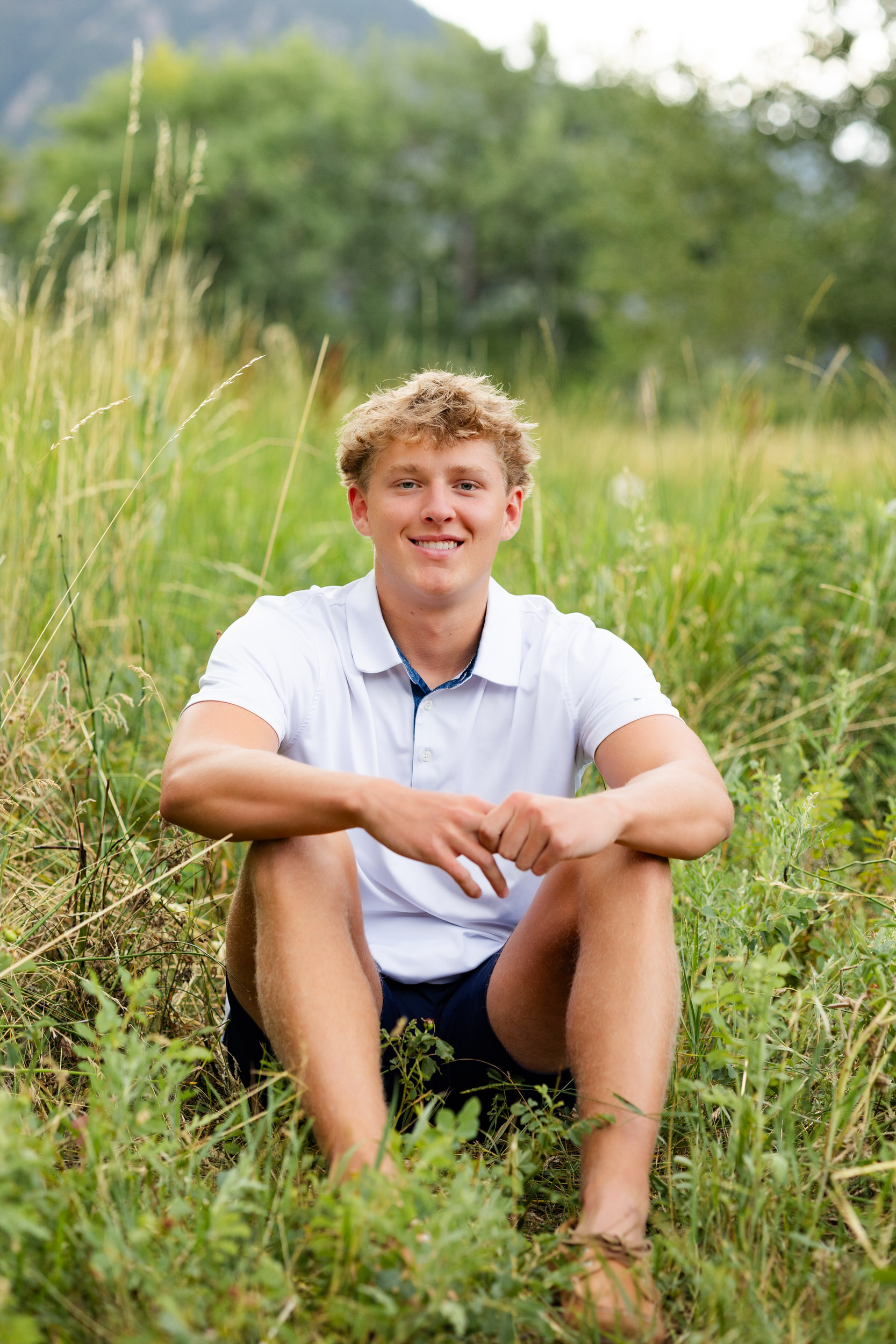 A young man sits in a field and smiles at the camera.