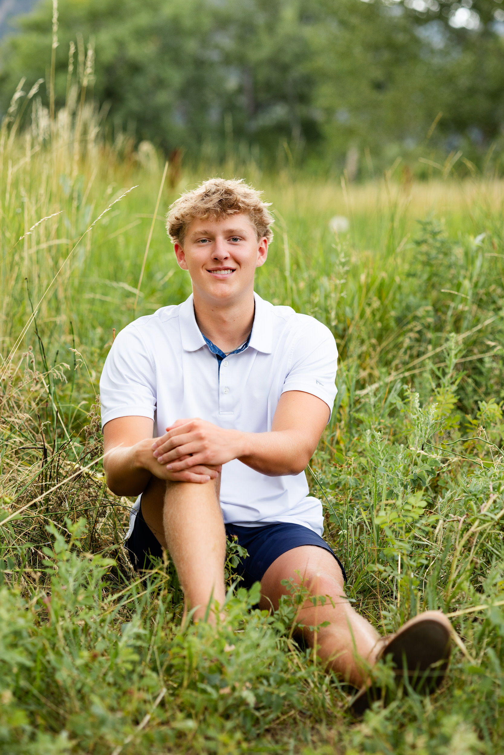 A young man sits in a field and smiles at the camera.