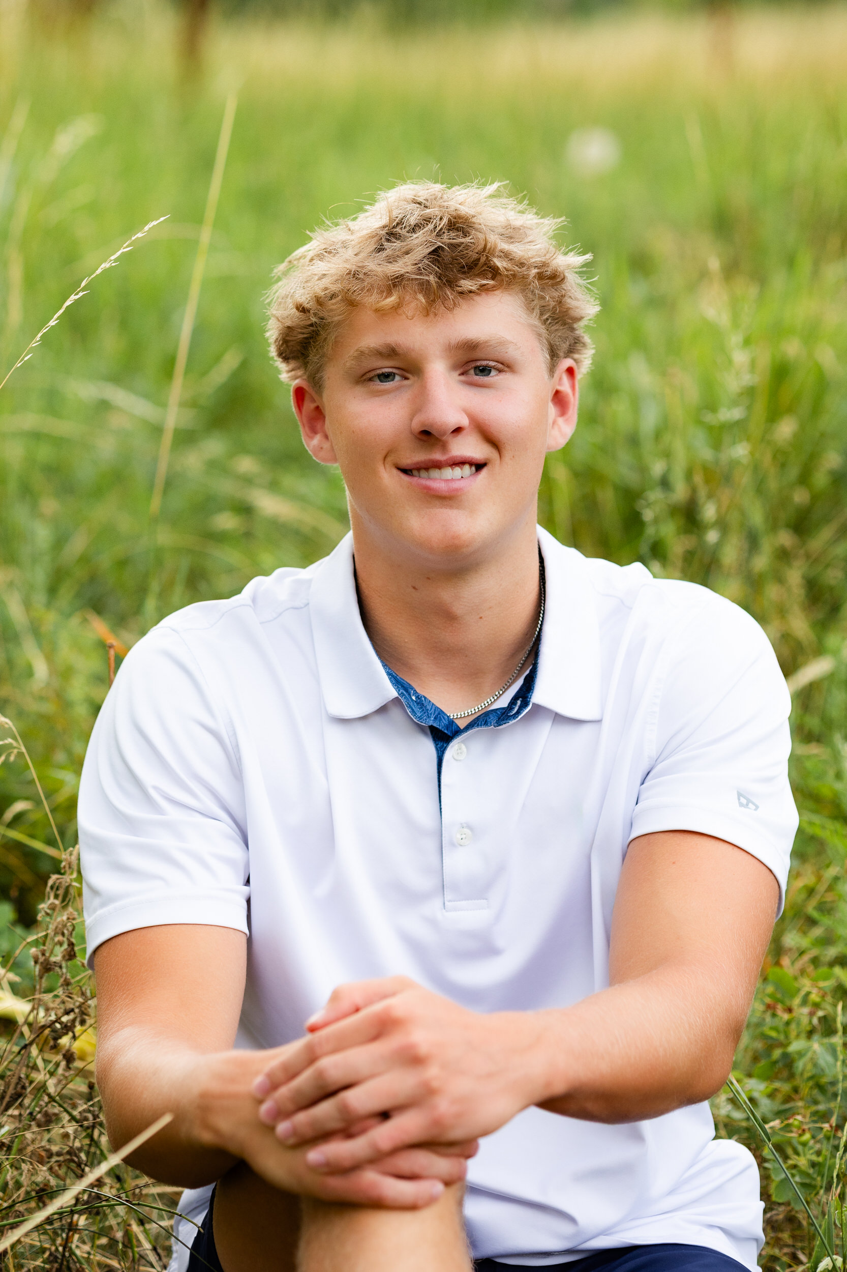 A young man sits in a field and smiles at the camera.