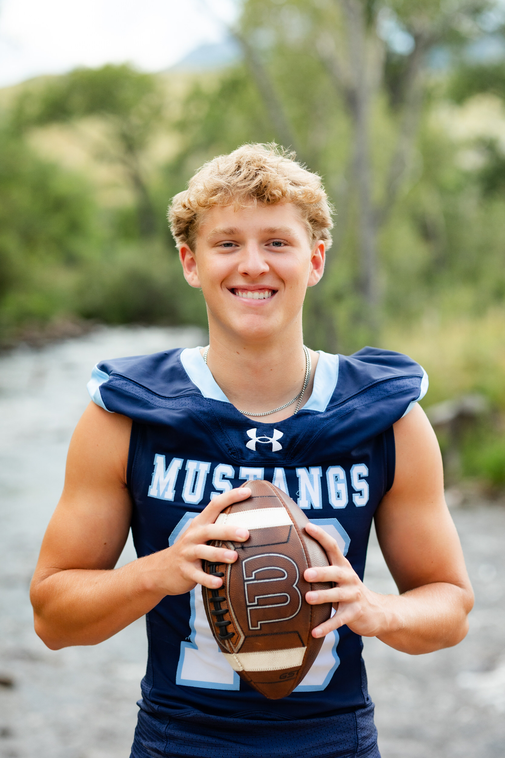 A young man in a school football jersey holds a football and smiles at the camera.