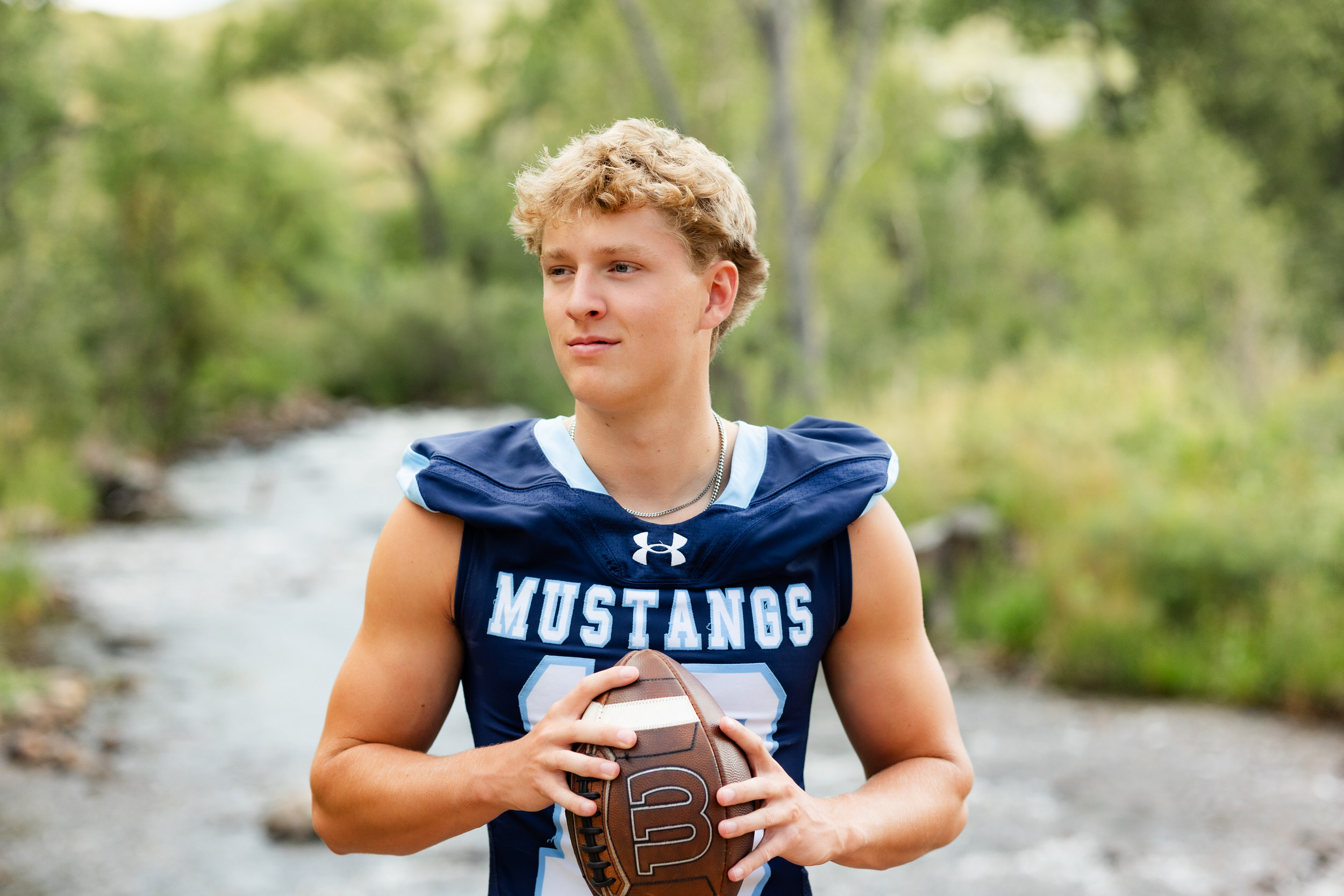 A young man in a school football jersey holds a football and looks off camera.