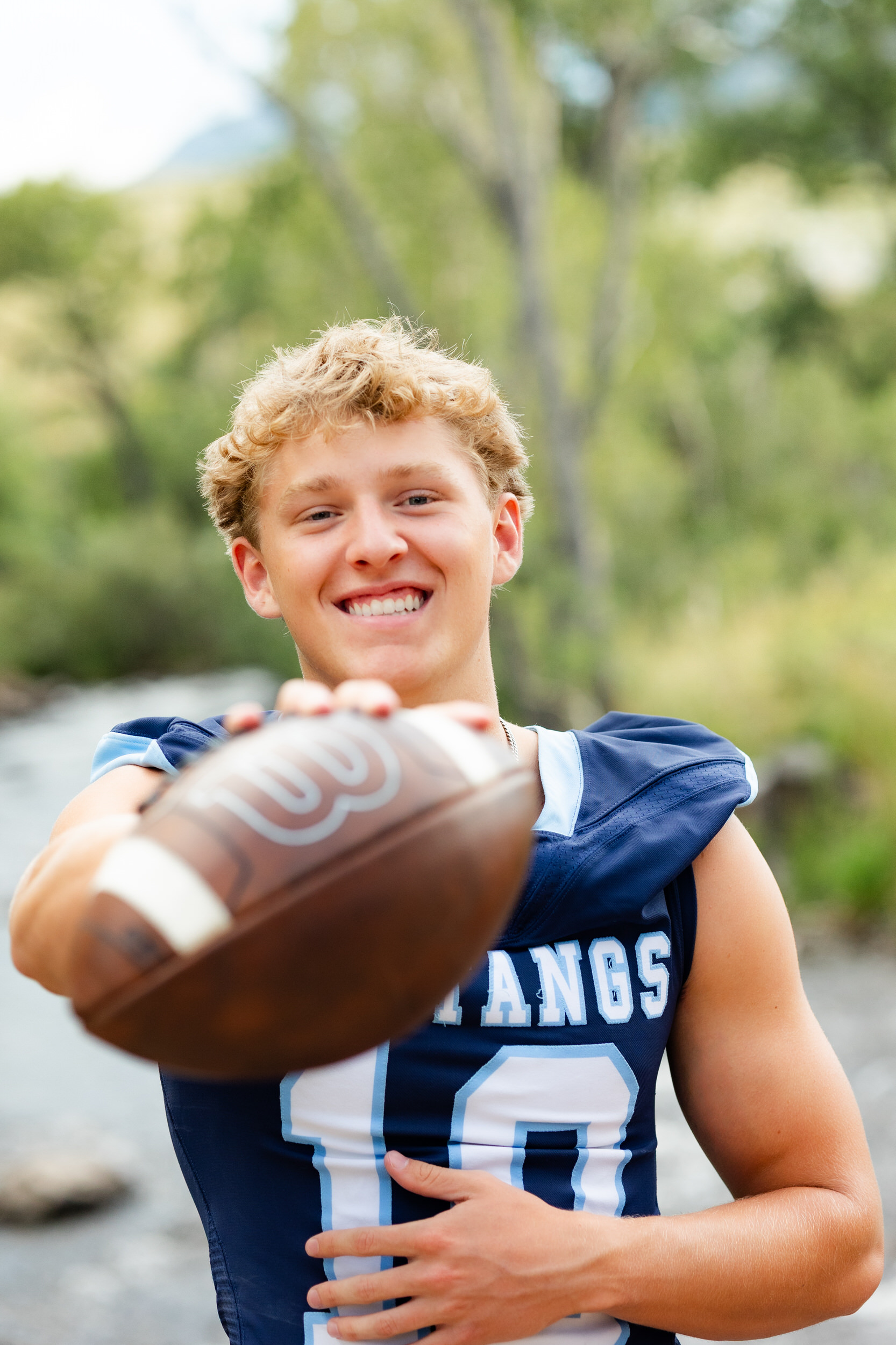 A young man in a school football jersey holds up a football toward the camera and smiles.