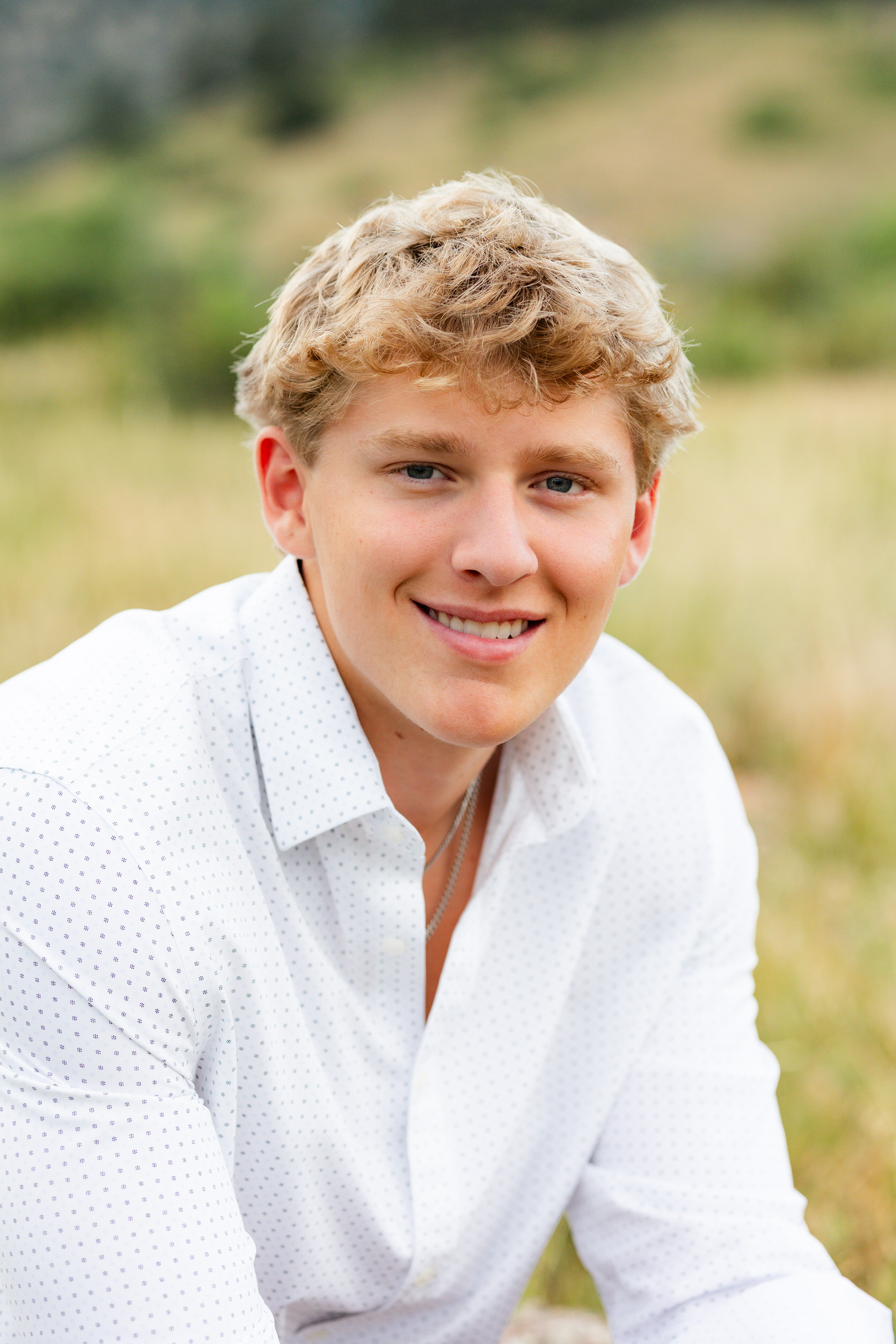 A young man sits in a field and smiles at the camera.