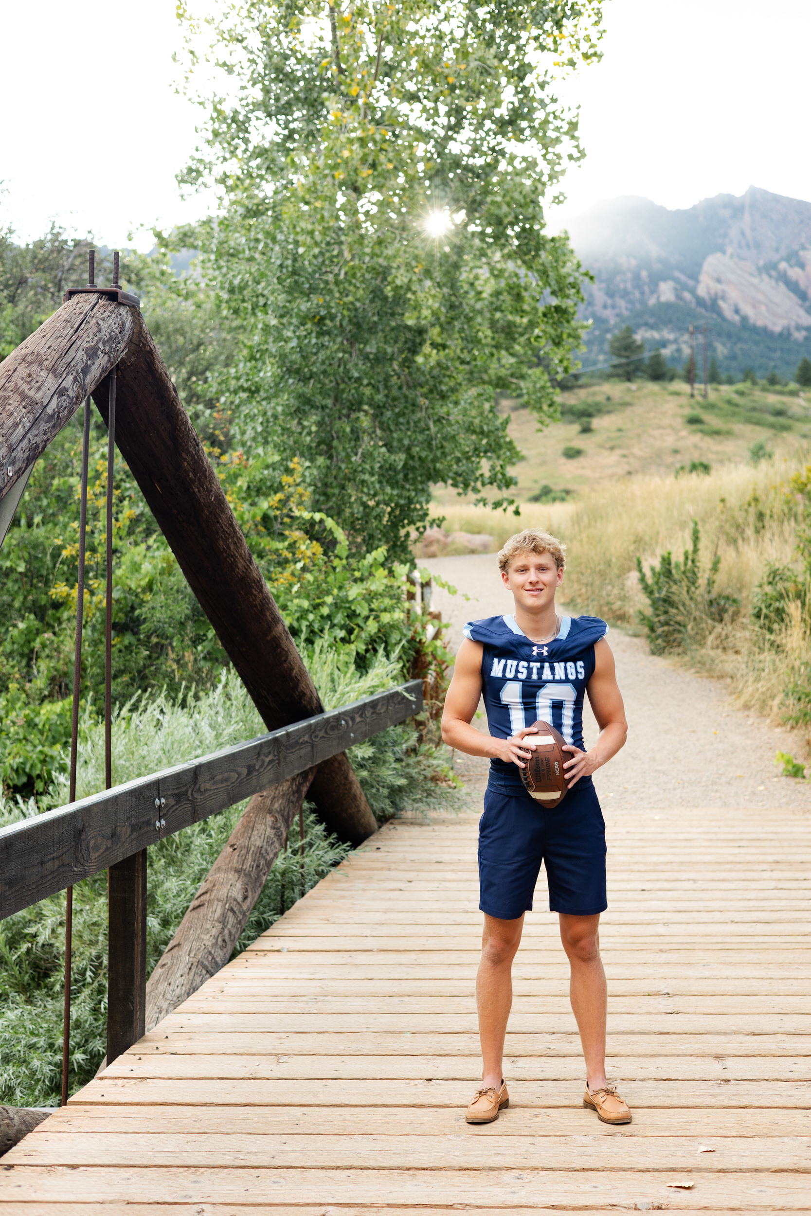 A young man in a school football jersey stands on a bridge in front of the mountains and smiles at the camera.