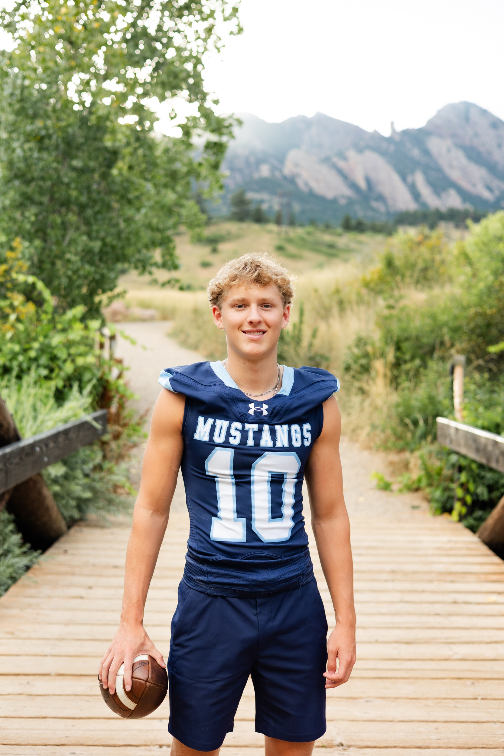 A young man in a school football jersey stands on a bridge in front of the mountains and smiles at the camera.
