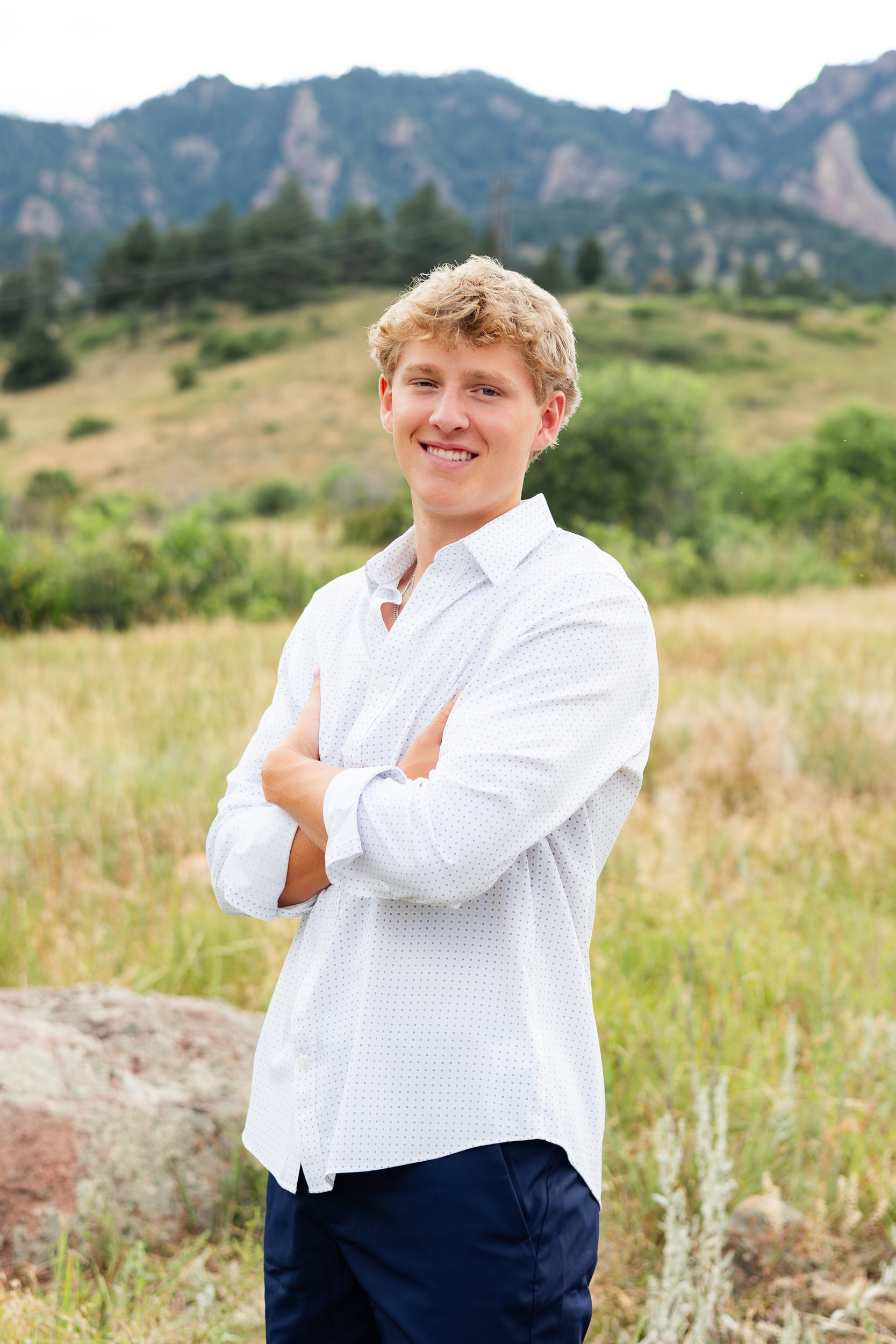 Young man stands in a mountain field with his arms crossed and smiles at the camera.