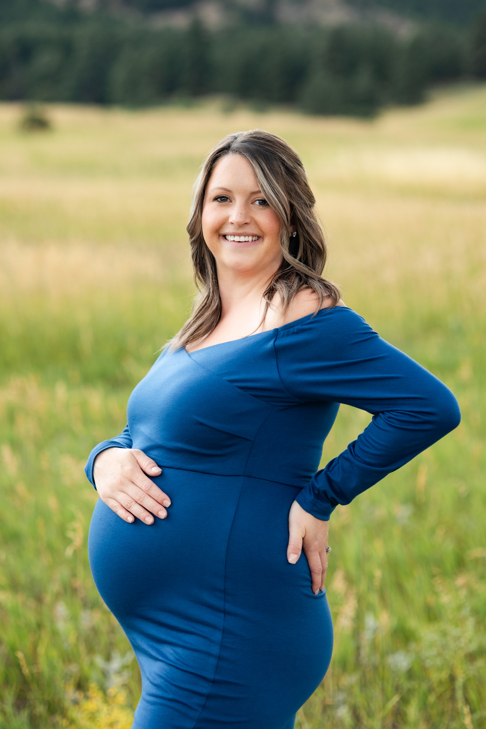 A pregnant woman in a blue dress smiles at the camera with one hand on her belly and one hand on her back.