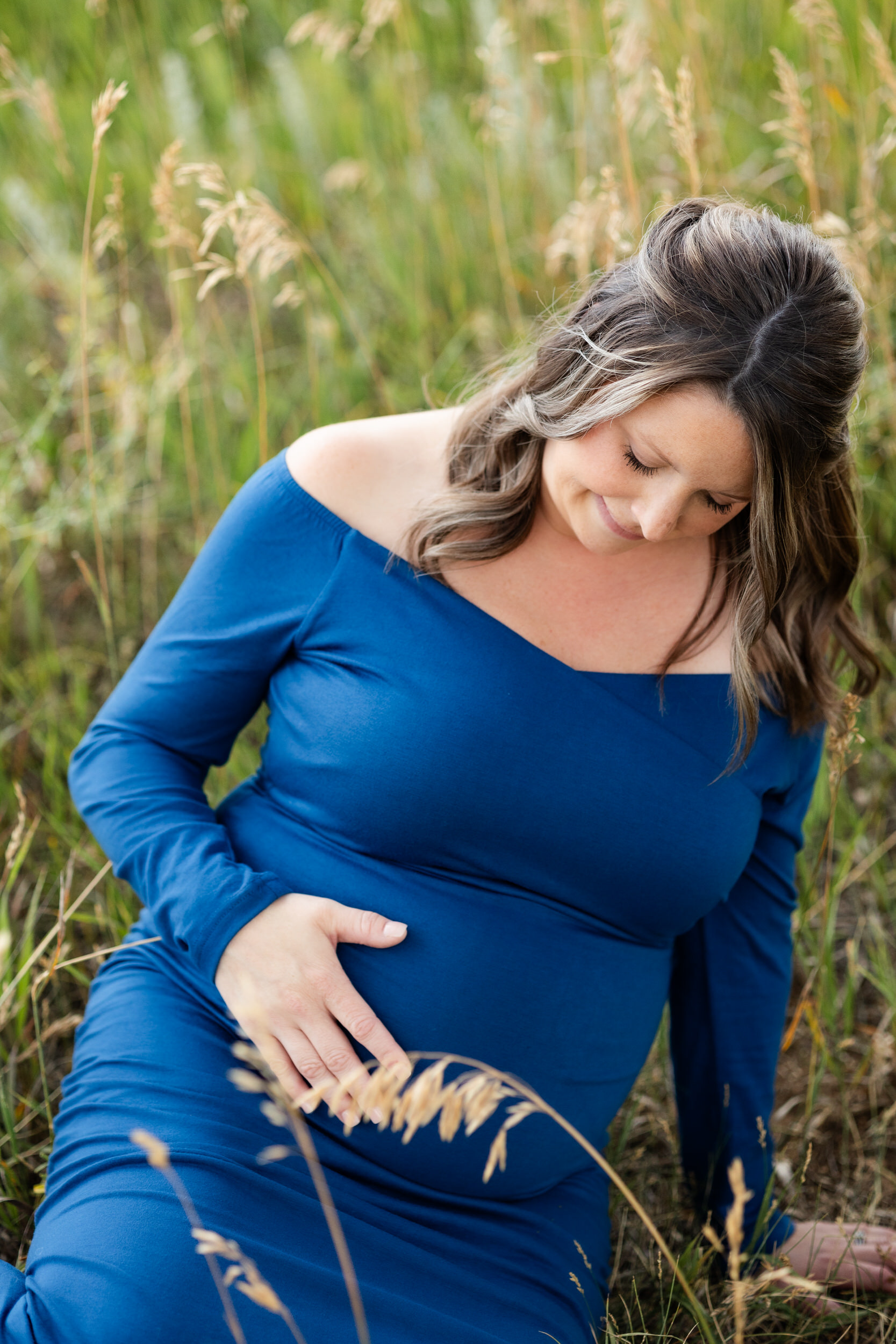 A pregnant woman sits in a field and smiles down at her bump.