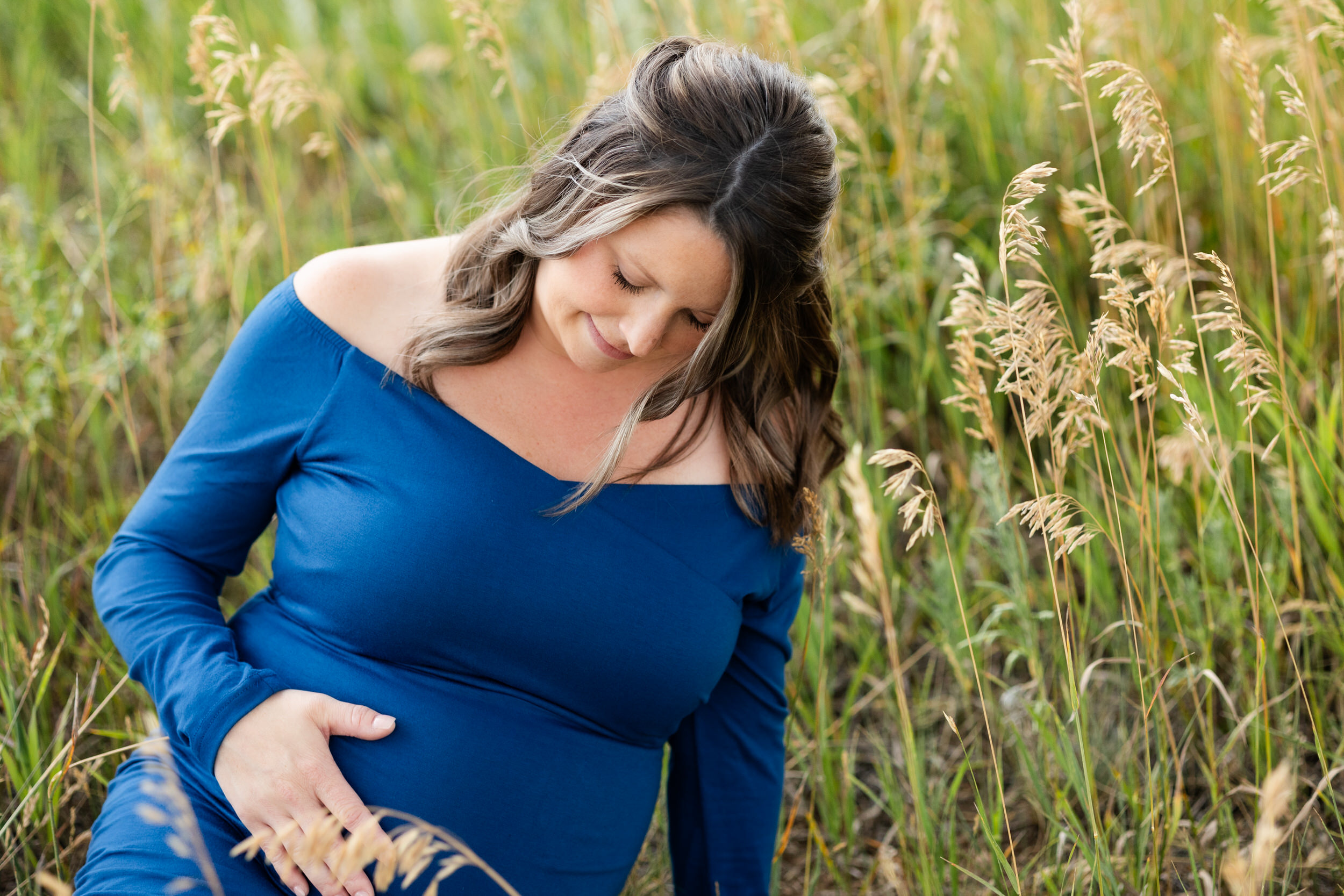 A pregnant woman sits in a field and smiles down at her bump.