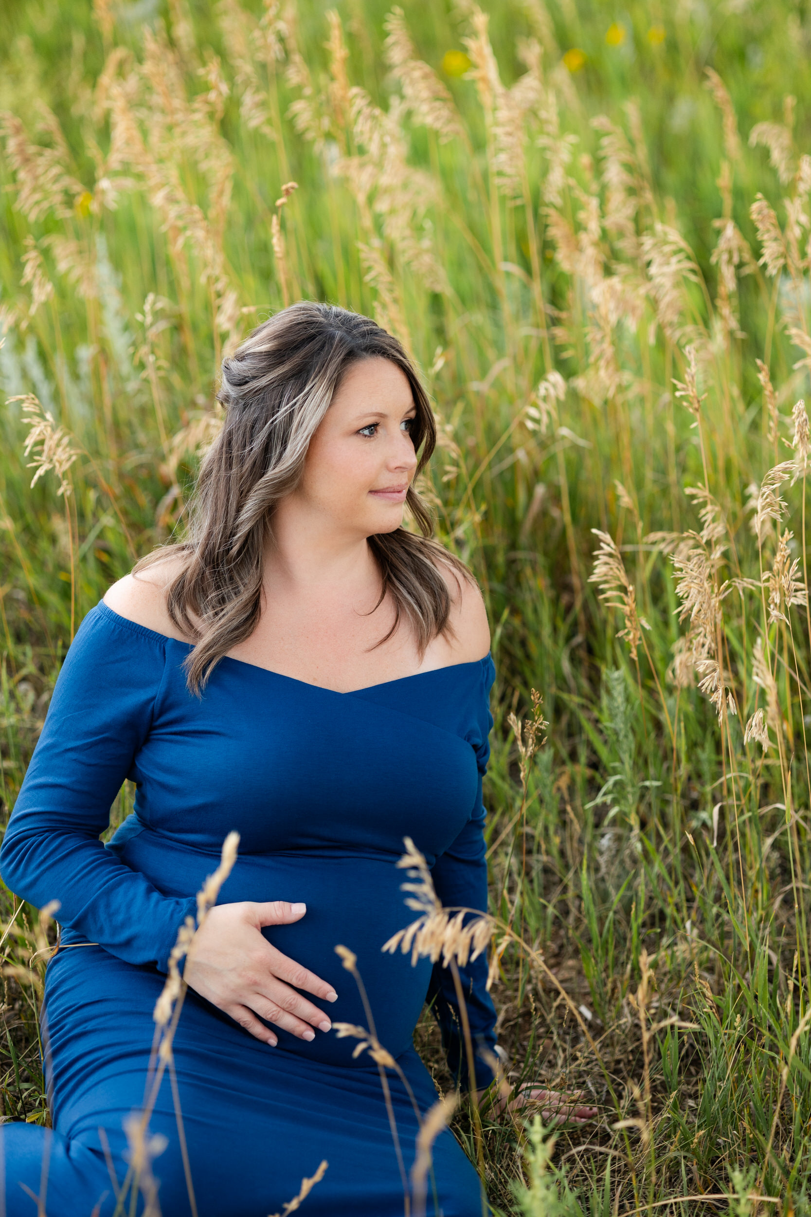 A pregnant woman sits in a field with one hand on her belly and looks off camera over her shoulder.