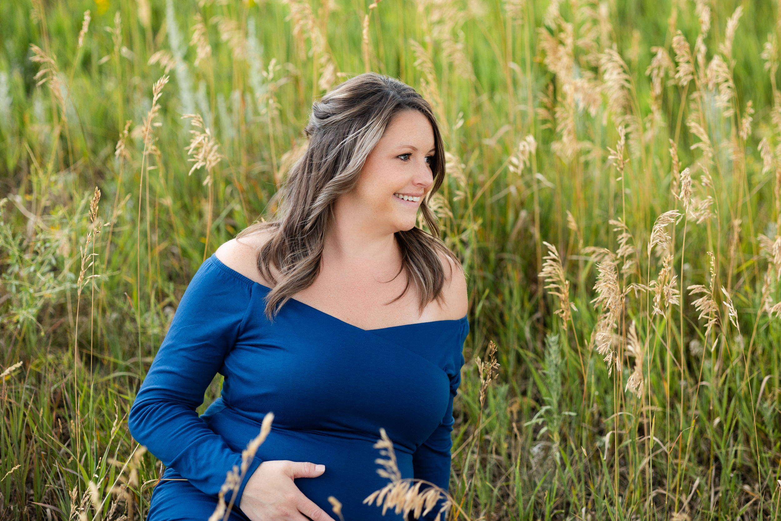 A pregnant woman sits in a field with one hand on her belly and smiles off camera.