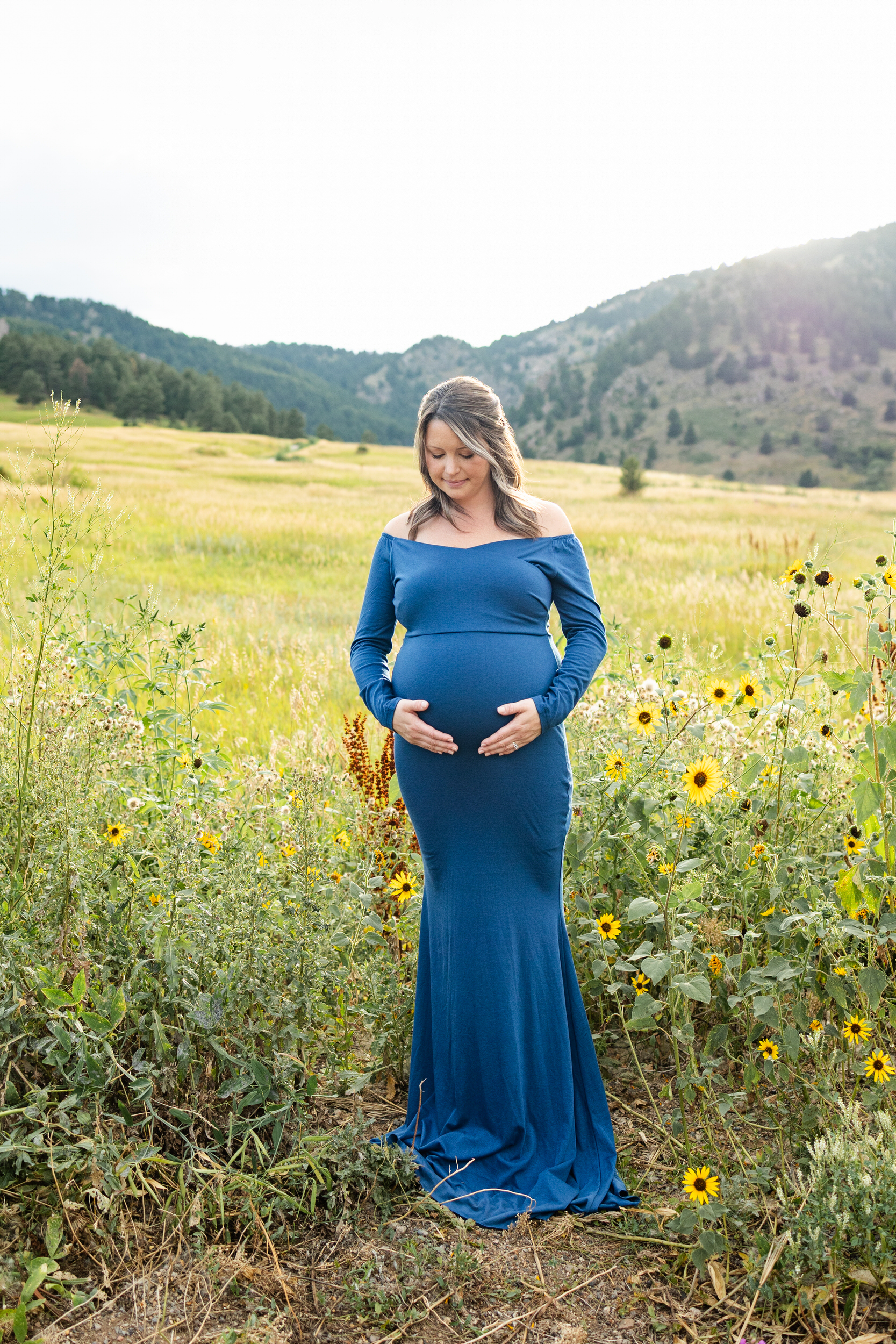 A pregnant woman in a blue dress stands in a field with both hands on her belly.
