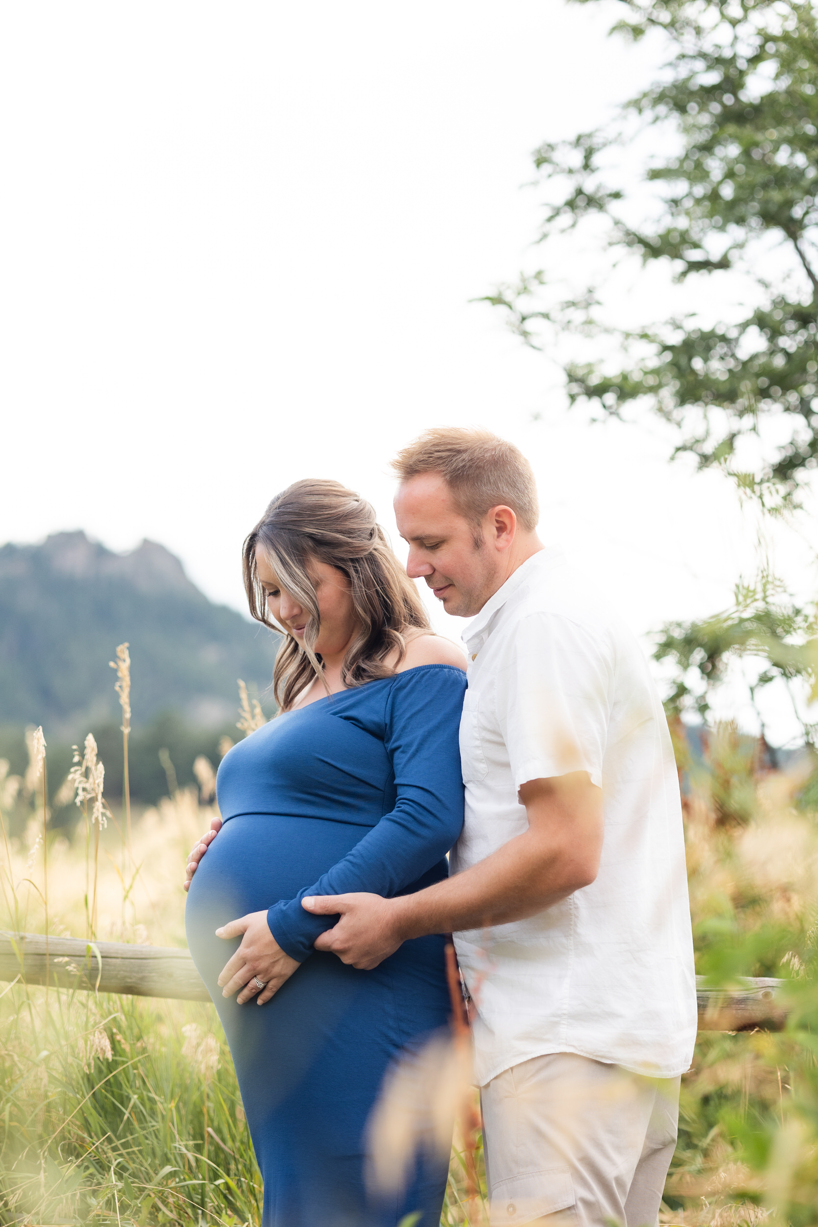 A pregnant woman stands with her back to her husband's chest and both of them have their hands on her pregnant belly smiling at the bump.