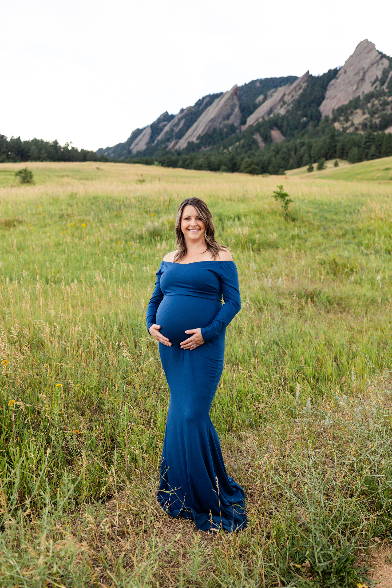 Pregnant woman in a blue dress stands in a field and smiles at the camera with both hands on her belly.