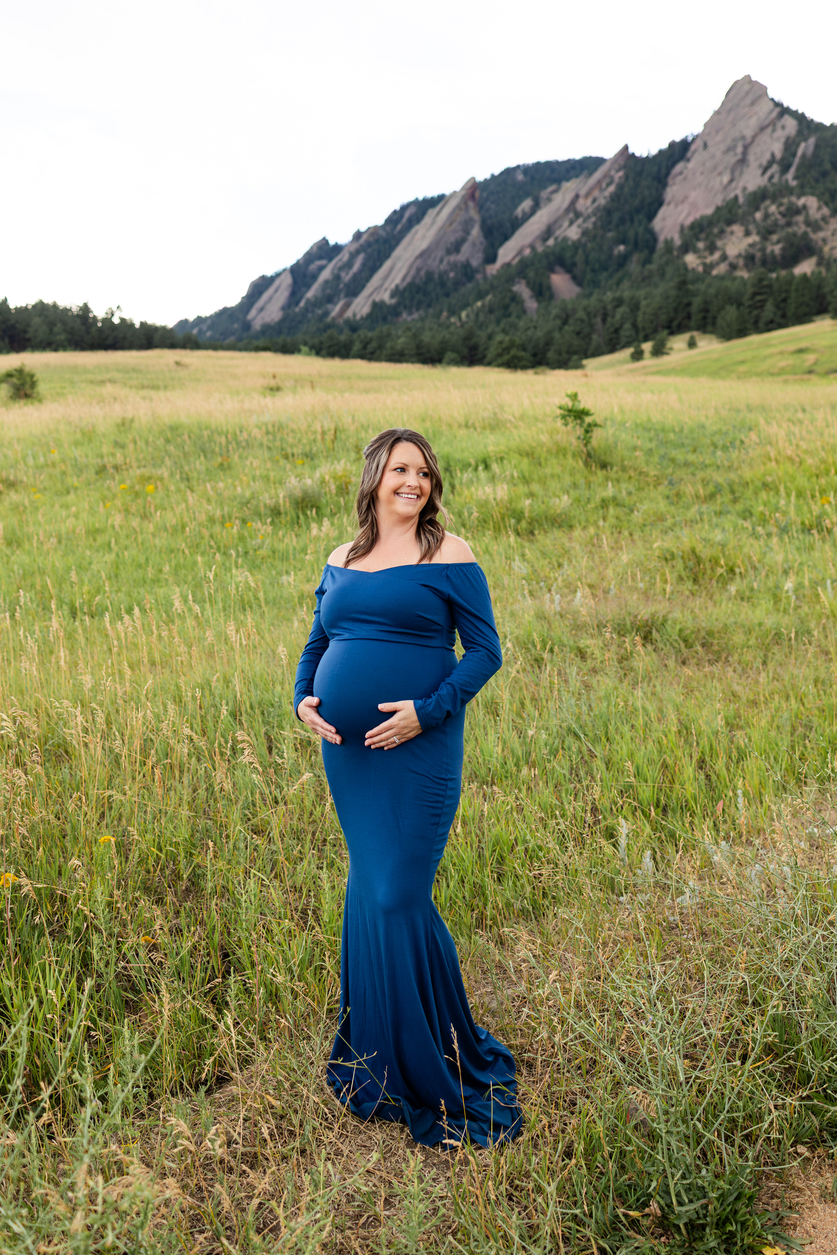 Pregnant woman in a blue dress stands in a field with both hands on her belly and smiles off camera.