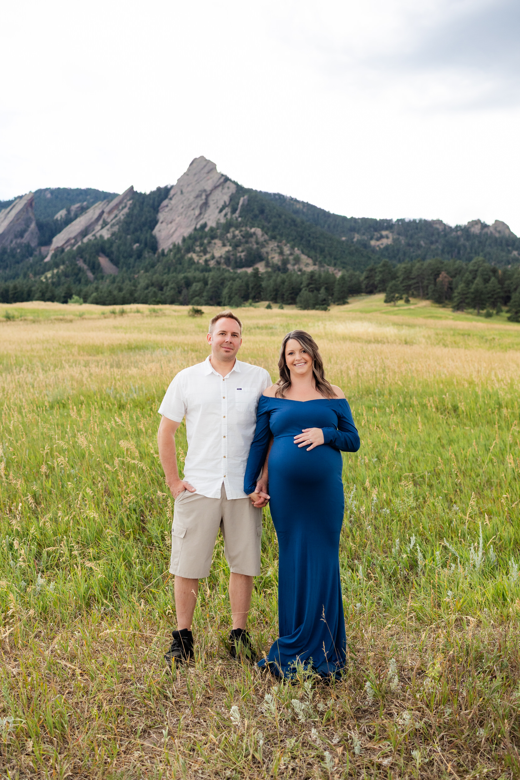 Husband and pregnant wife stand side by side smiling at the camera in a mountain field.