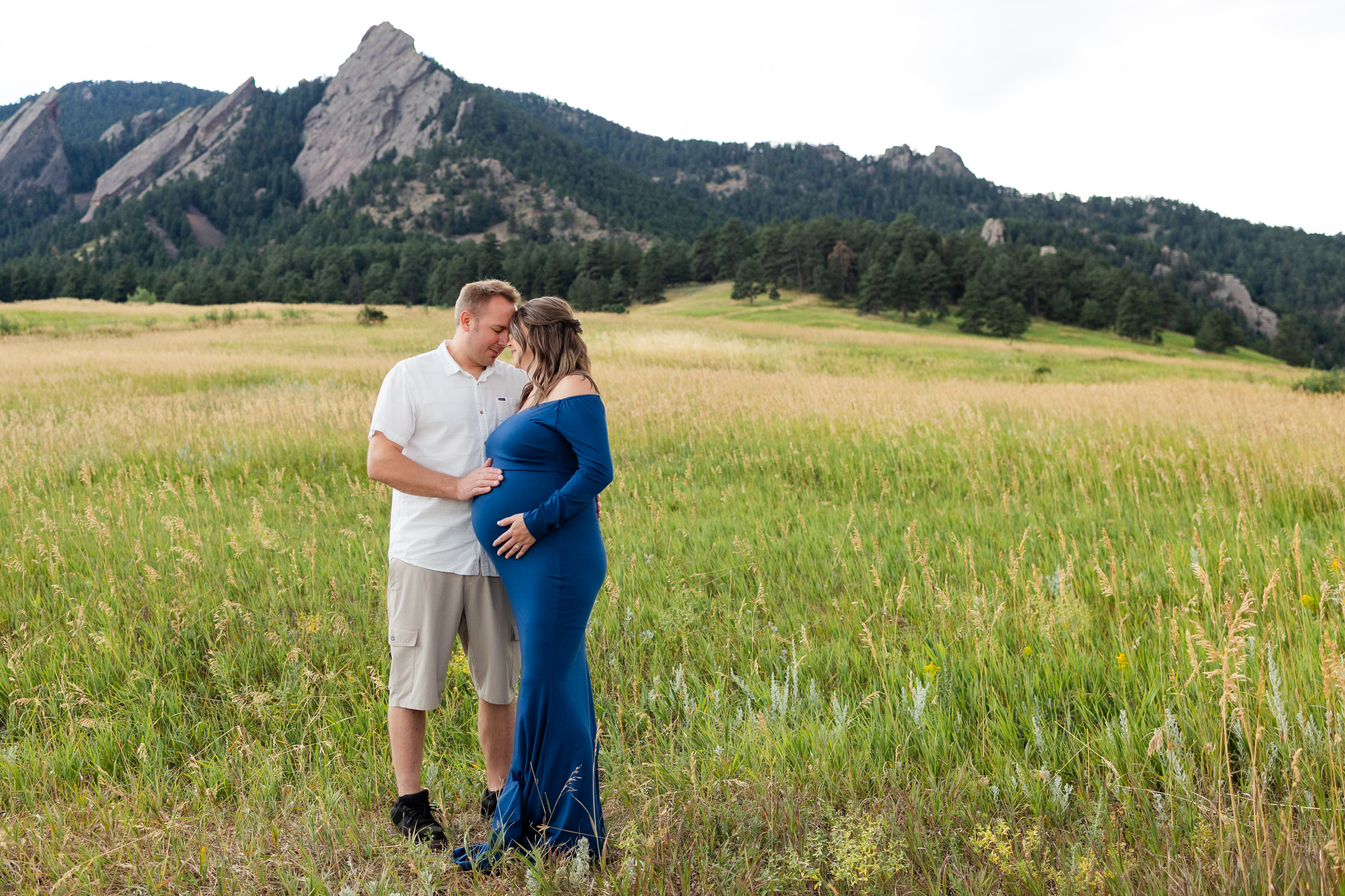 Husband and pregnant wife stand forehead to forehead in a mountain field.