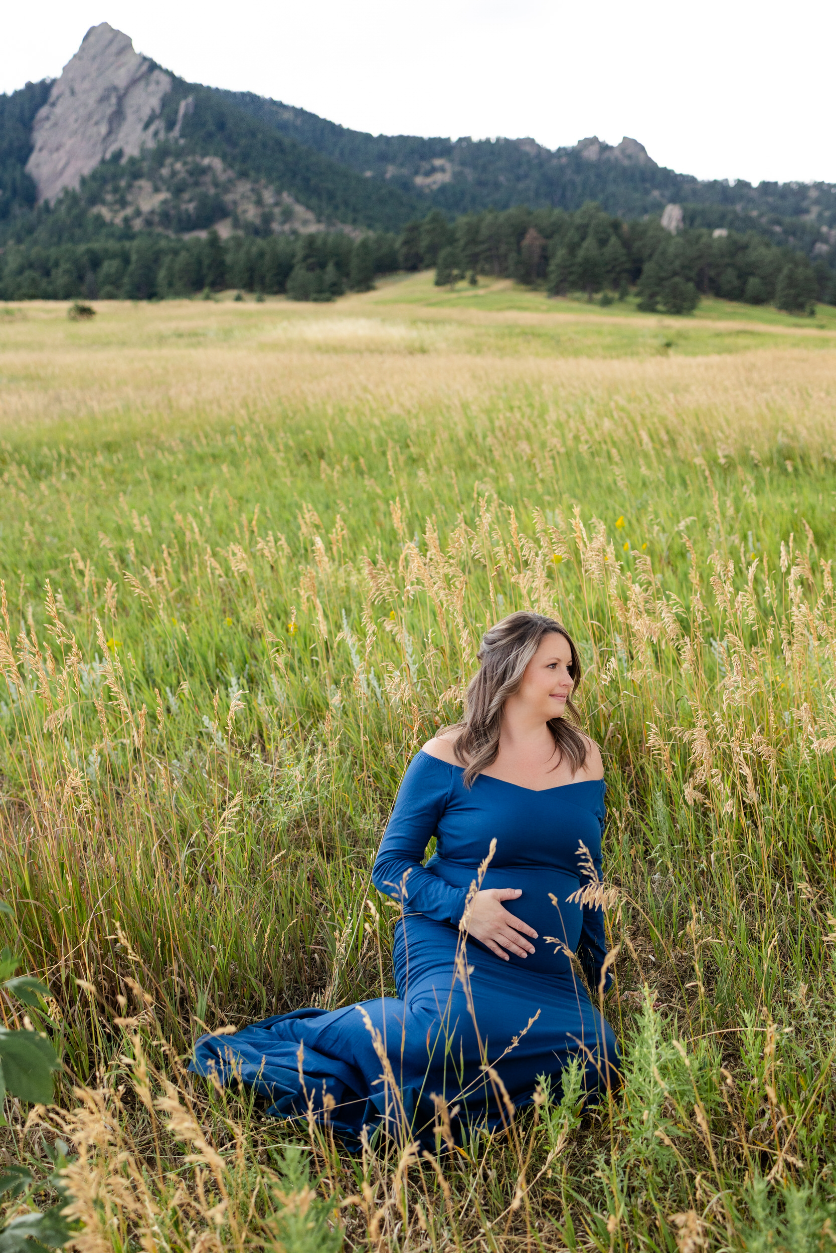 Pregnant woman in a blue dress sits in a mountain field with one hand on her belly smiling off camera.